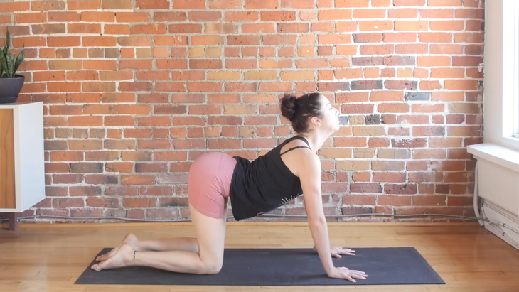 Woman in Cow Pose on a yoga mat placed on a hard wood floor with a brick wall background