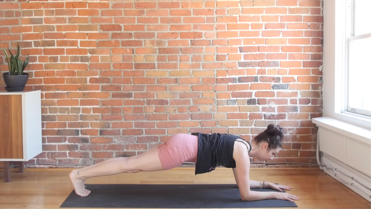 Forearm Plank on a yoga mat placed on a hardwood floor with a brick wall backdrop