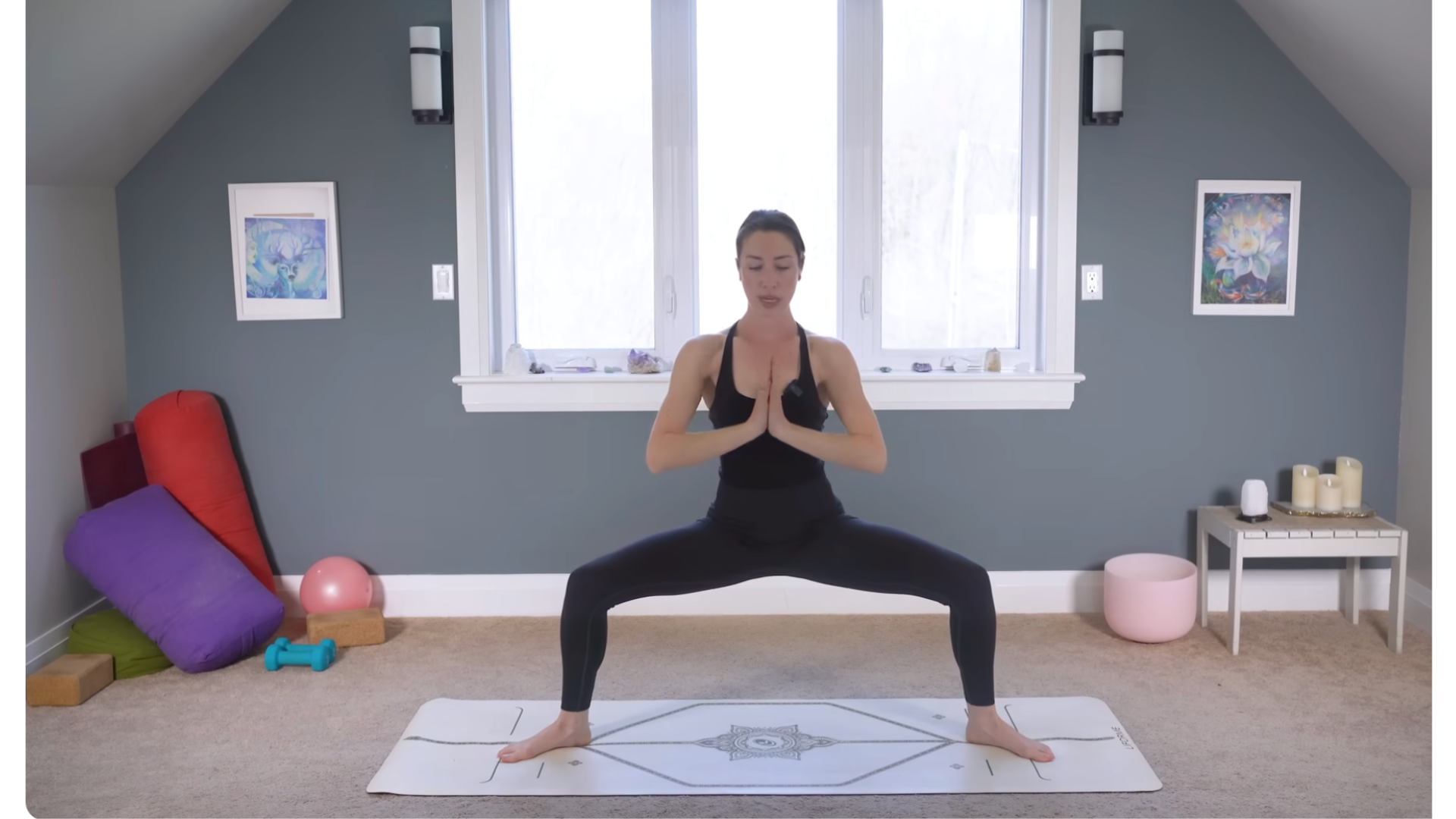Yoga teacher practicing Goddess Pose on a mat with her knees bent and her hands at her chest