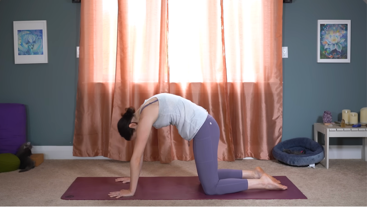Woman kneeling on a yoga mat with her back rounded in Cat Pose while practicing full-body stretches