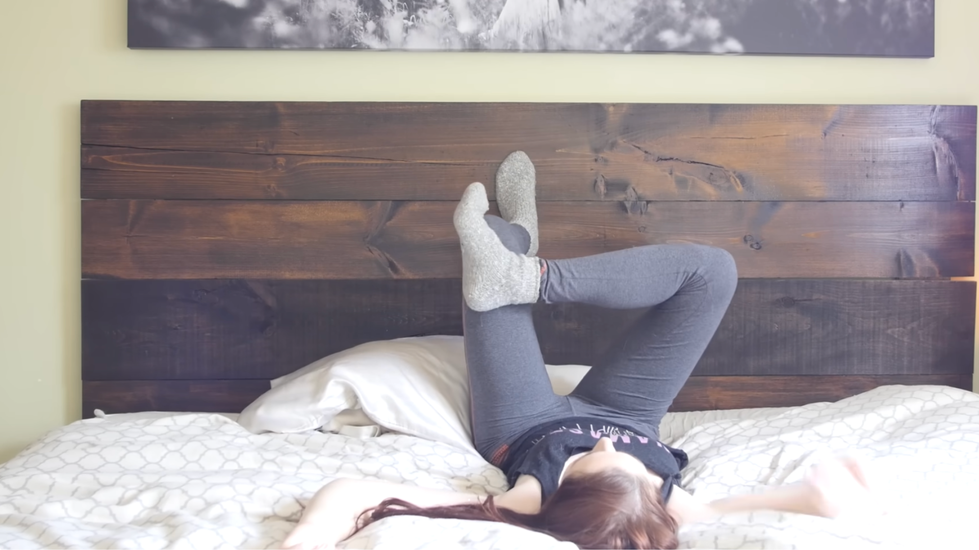 Yoga teacher lying in bed with one foot on the headboard and the other in a reclined figure-4 position