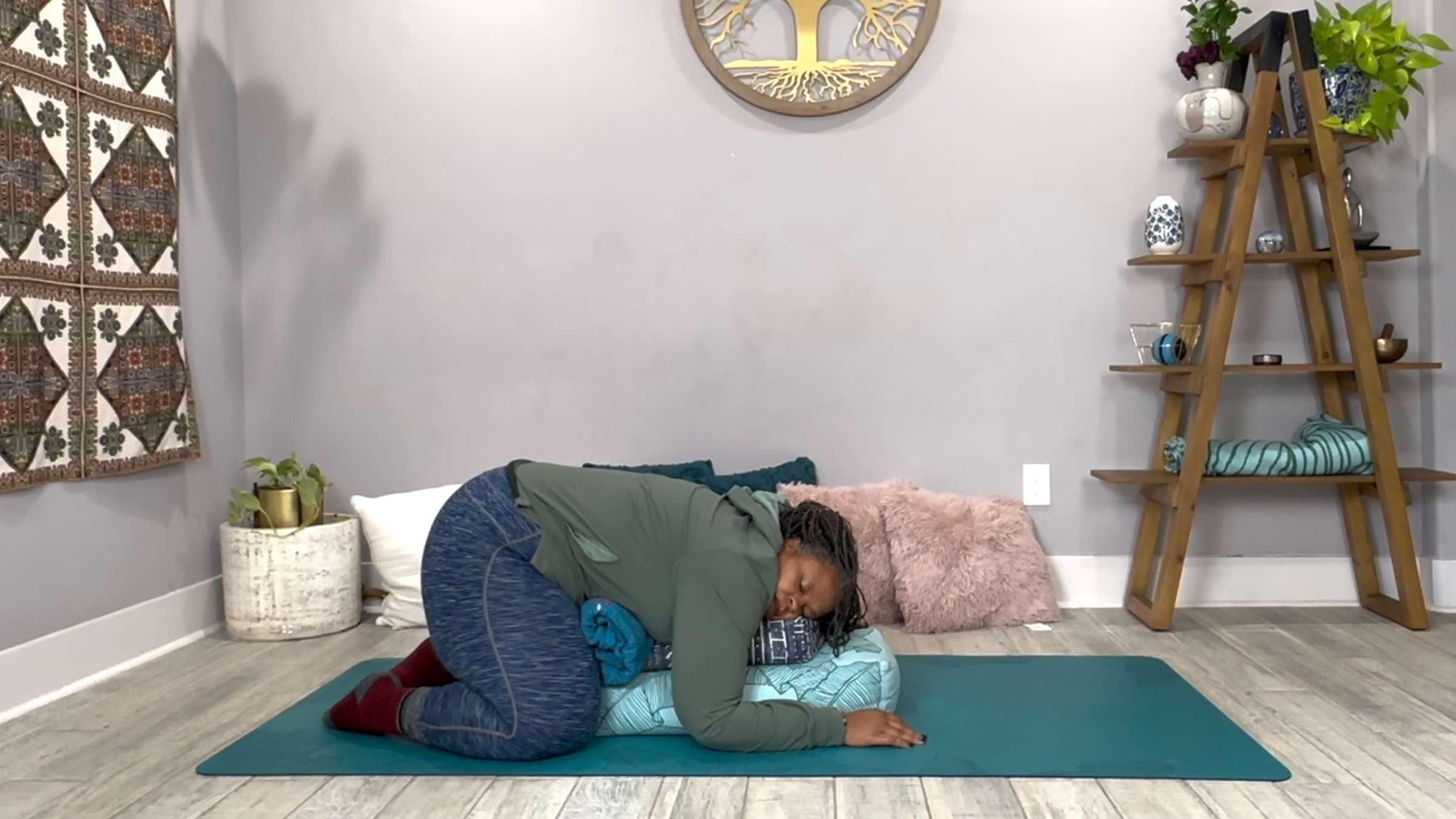 A yoga teacher in a Restorative Yoga pose, on her knees with her torso laid over a pile of pillows. 