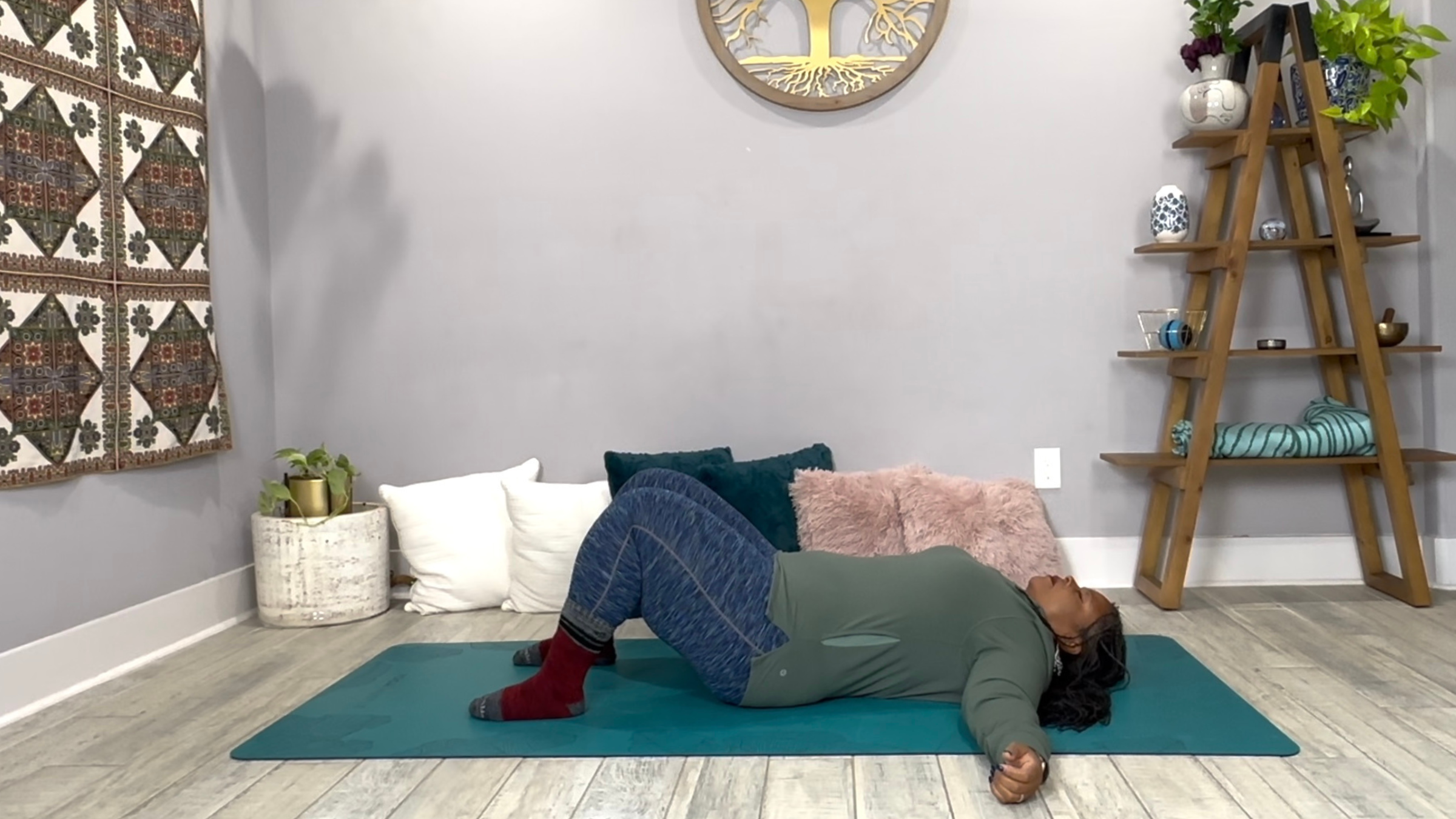 A yoga teacher practicing a Restorative Yoga pose, lying flat on her back with knees bent and arms out. 