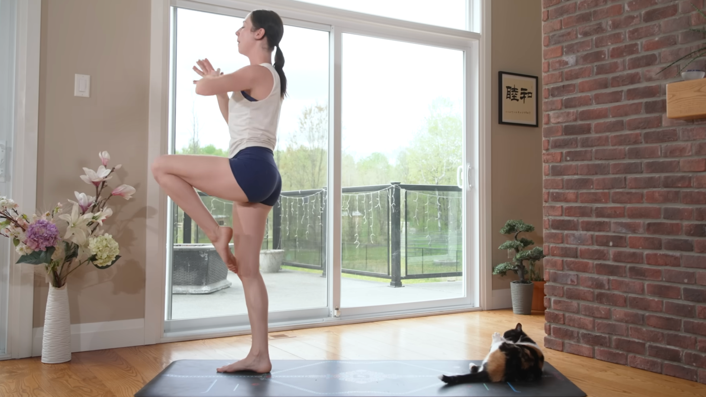 woman stands in position to move into warrior 3 pose on yoga mat in home with cat lying on mat behind her