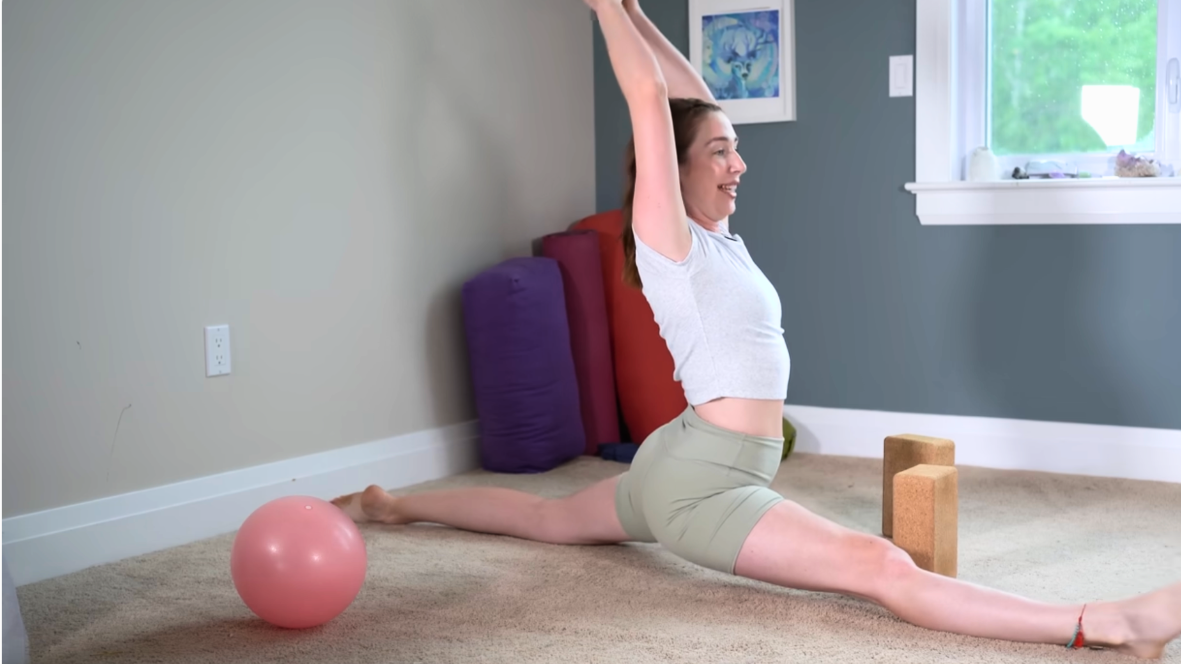 yoga instructor in splits position with arms raised on carpeted floor with blocks and ball nearby
