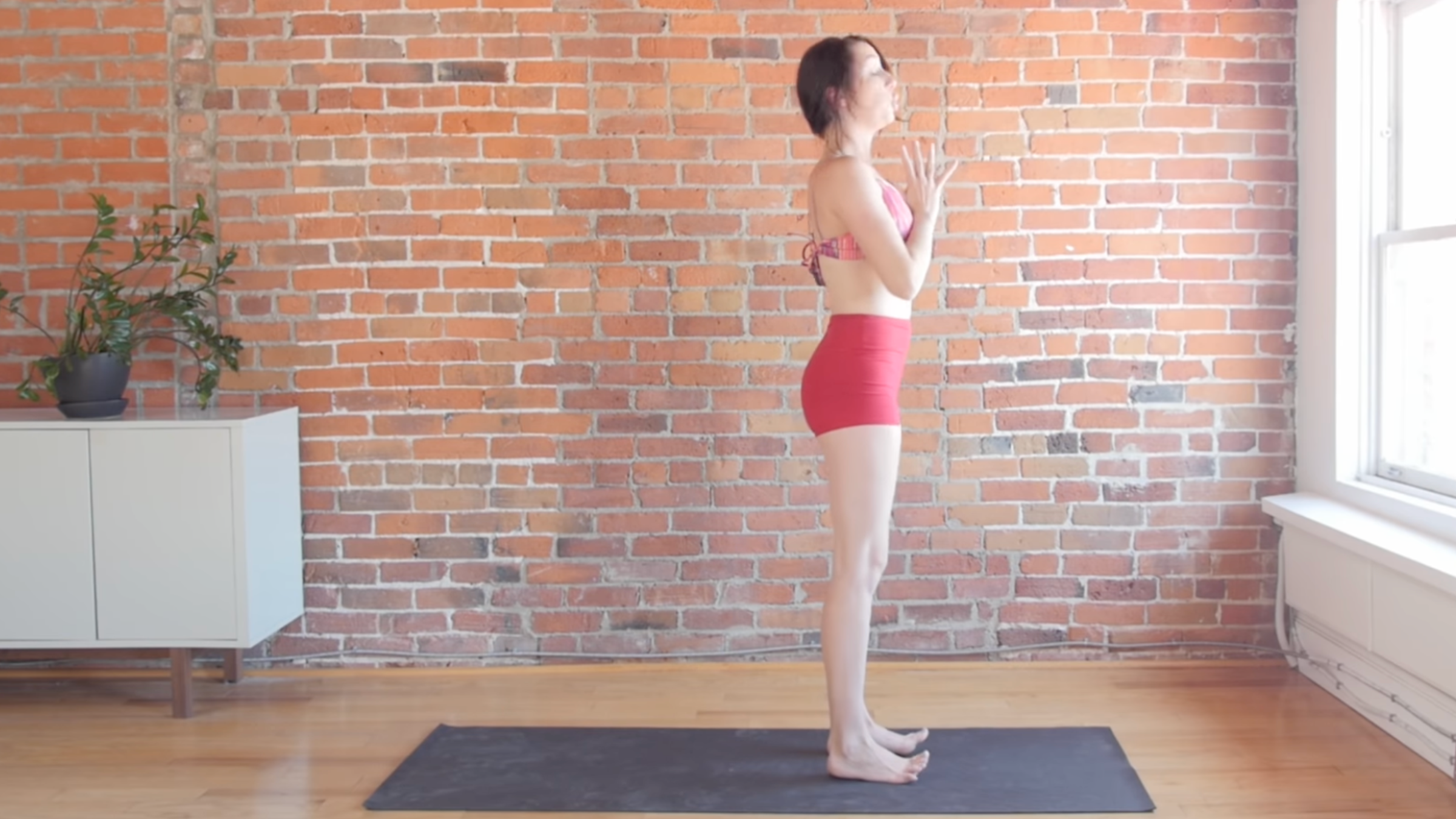 woman in standing sun salutations positions with hands together at chest on black yoga mat in front of brick wall
