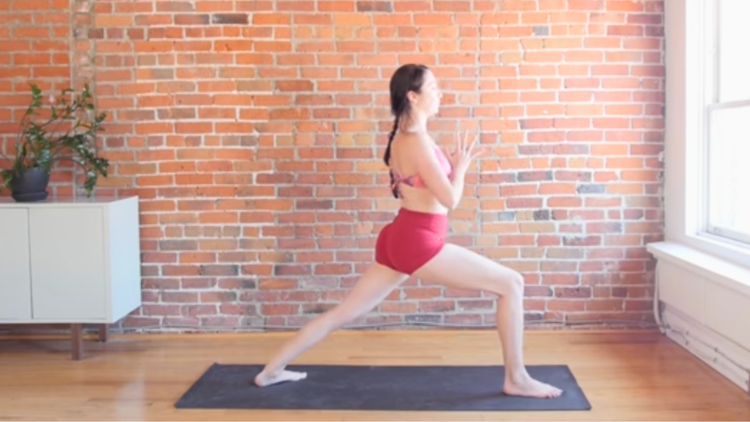 woman in warrior 1 pose with hands together at chest on black yoga mat in front of brick wall