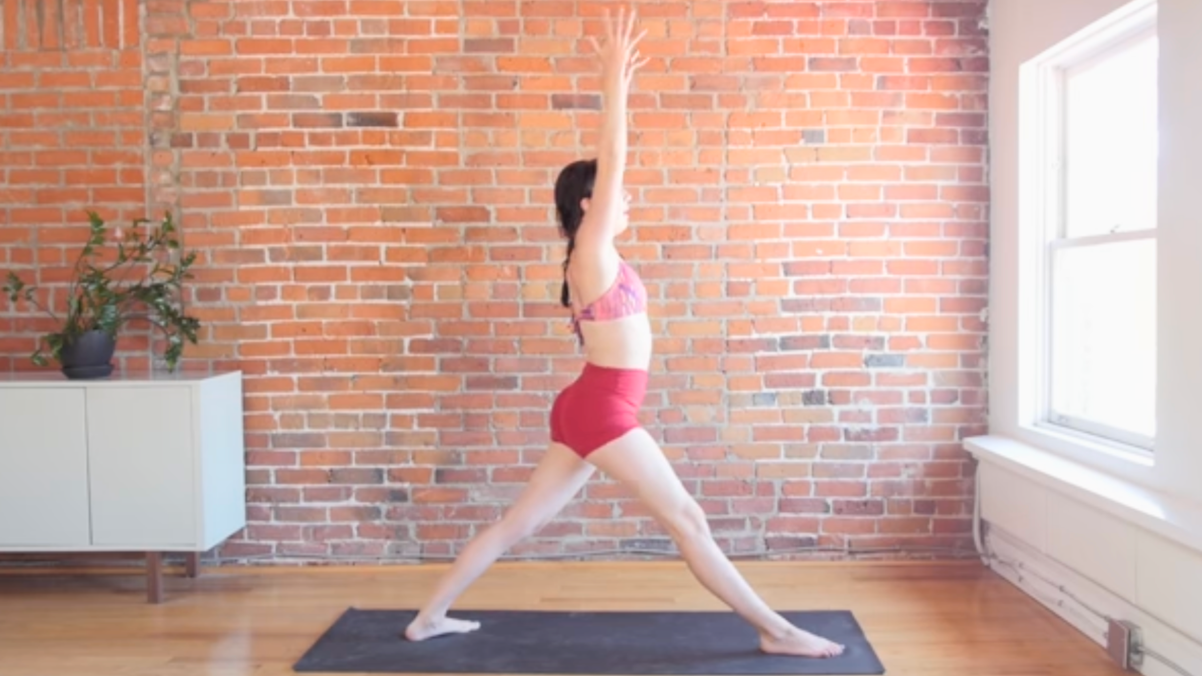 woman standing on black yoga mat with arms raised and feet split apart, in front of brick wall