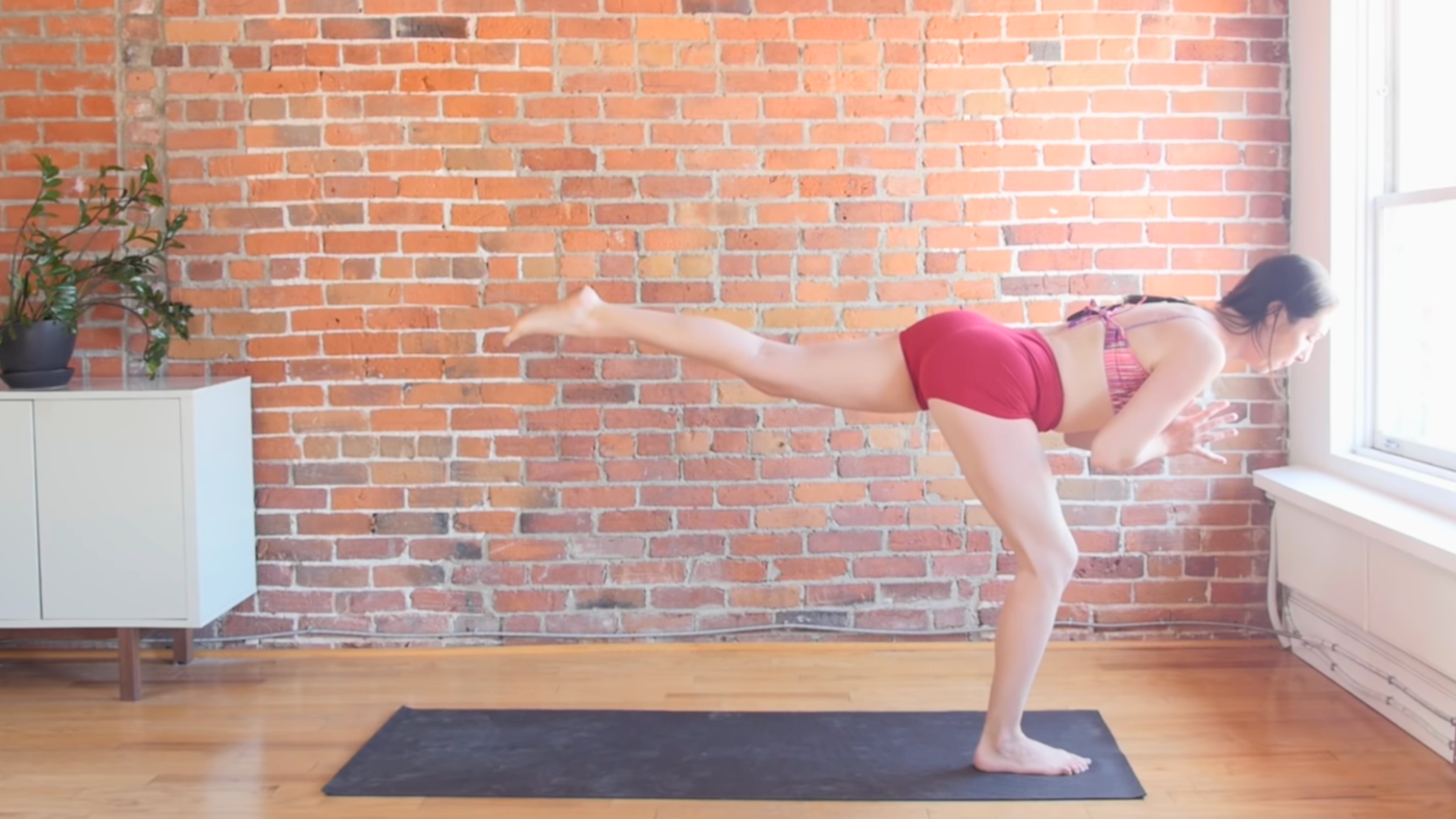 woman standing on yoga mat on right leg with slightly bent knee with torso and left leg parallel to the floor and hands together at chest