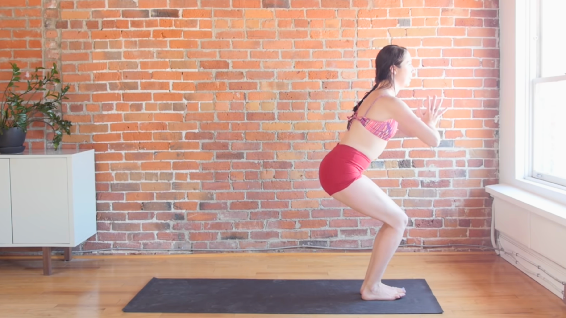 woman in chair pose with hands together in front of her on black yoga mat in front of brick wall