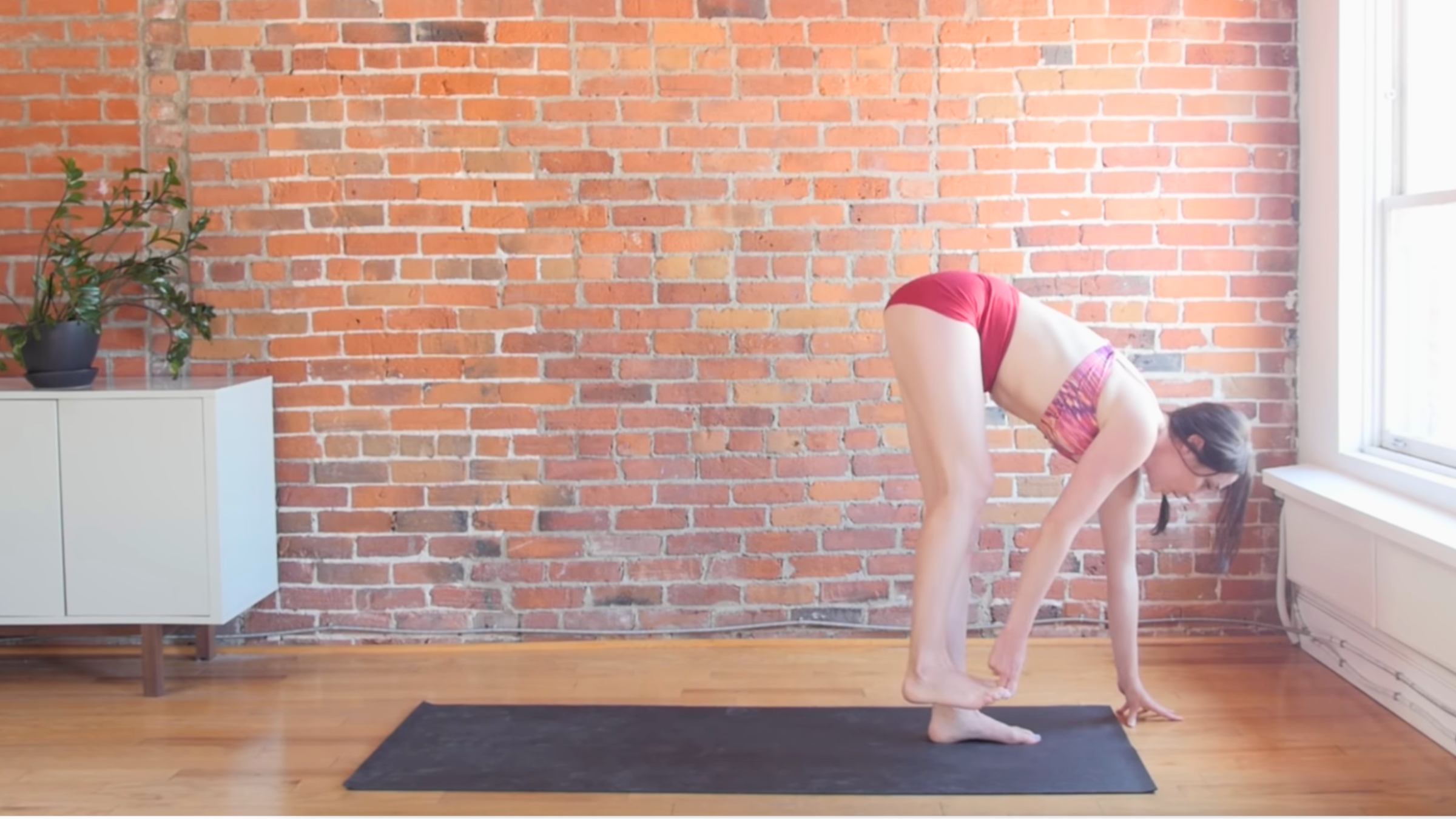 woman in balancing standing forward bend with fingers around right big toe on lifted right foot and left hand on ground on black yoga mat