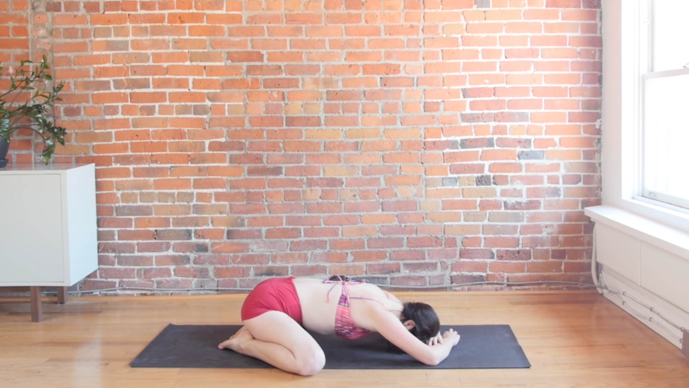 woman in child's pose with toes together and knees apart on black yoga mat in front of brick wall