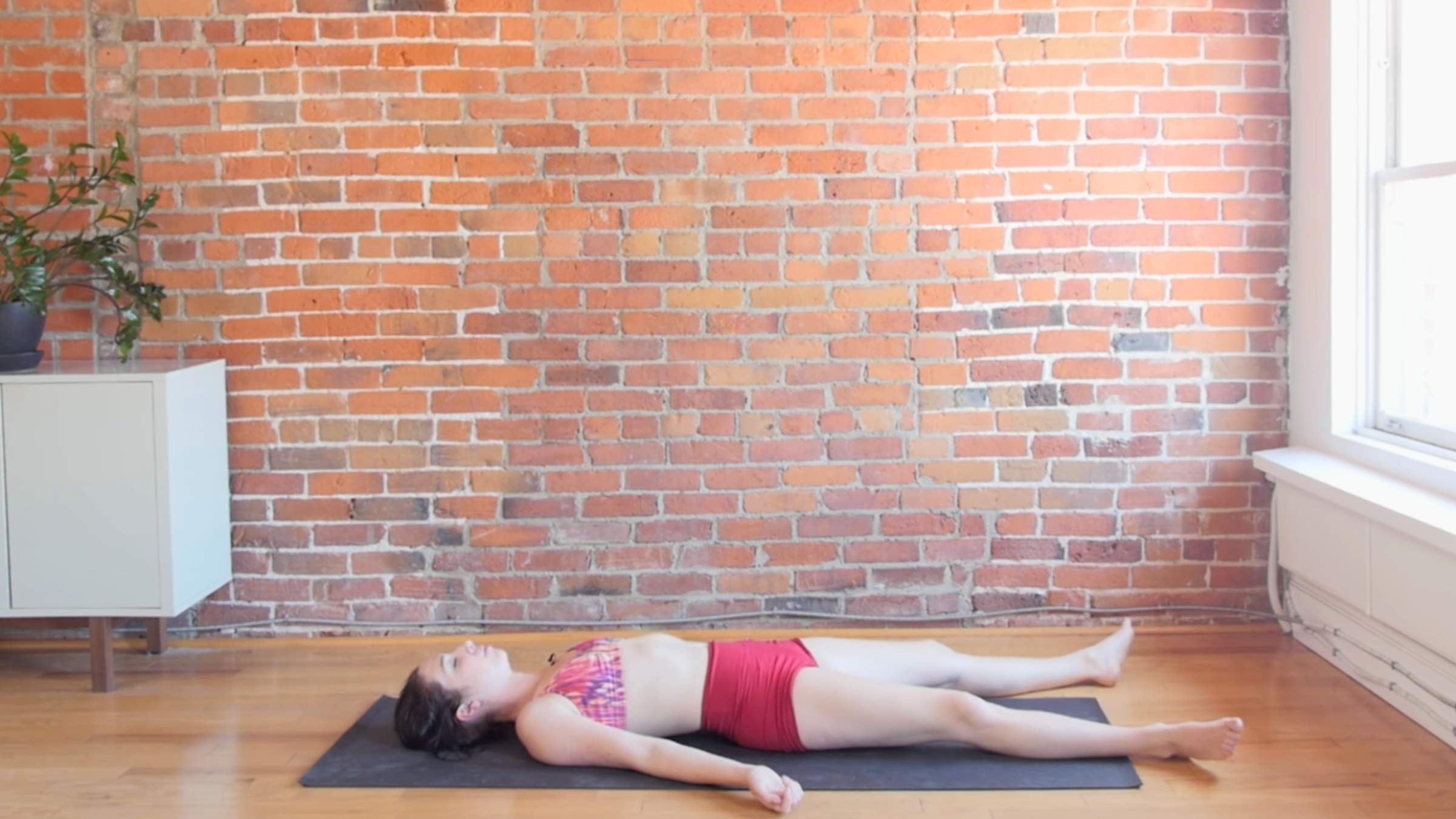 woman in corpse pose on black yoga mat in front of brick wall