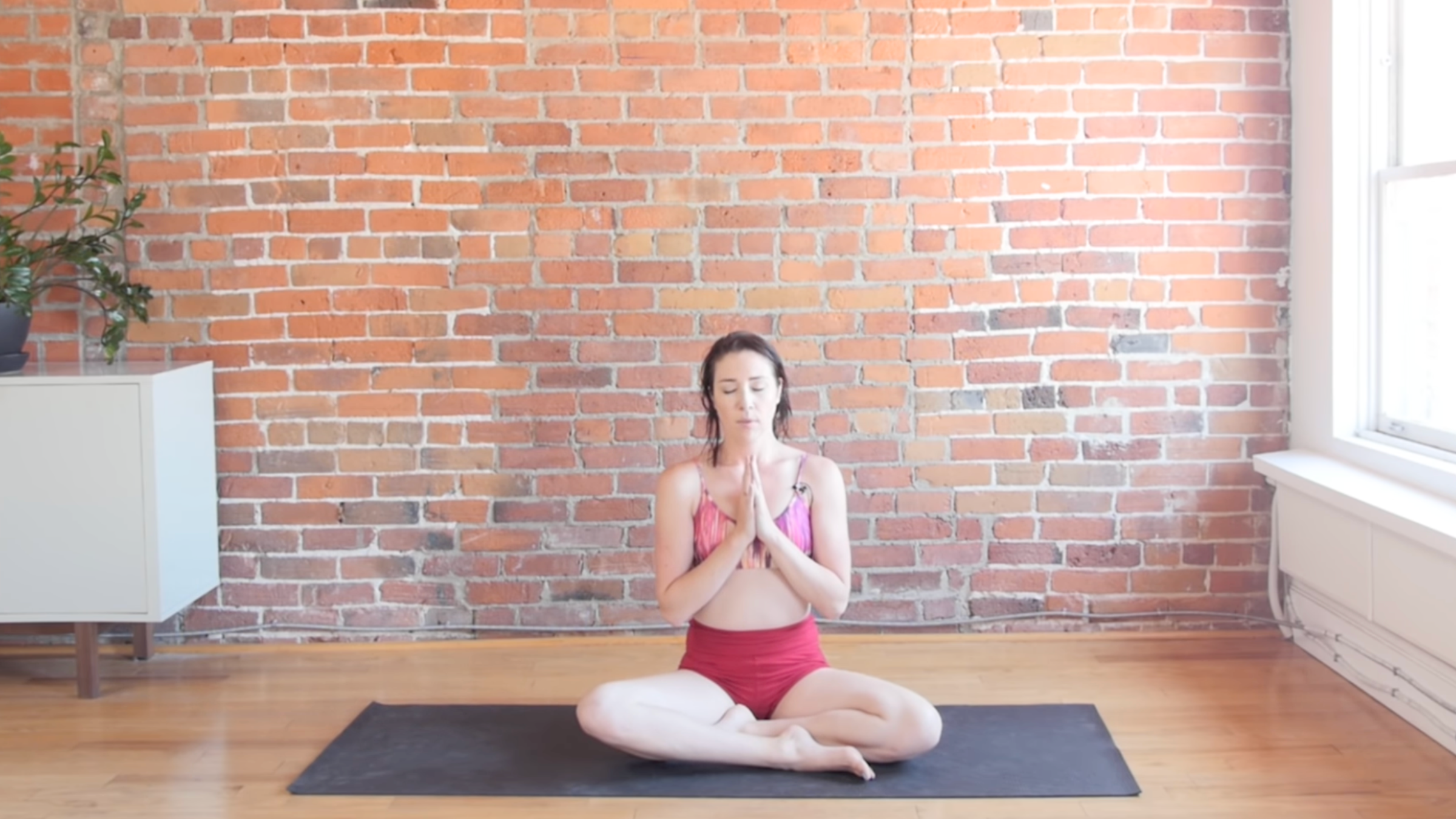 woman in cross-legged seat with hands together at heart on black yoga mat in front of brick wall
