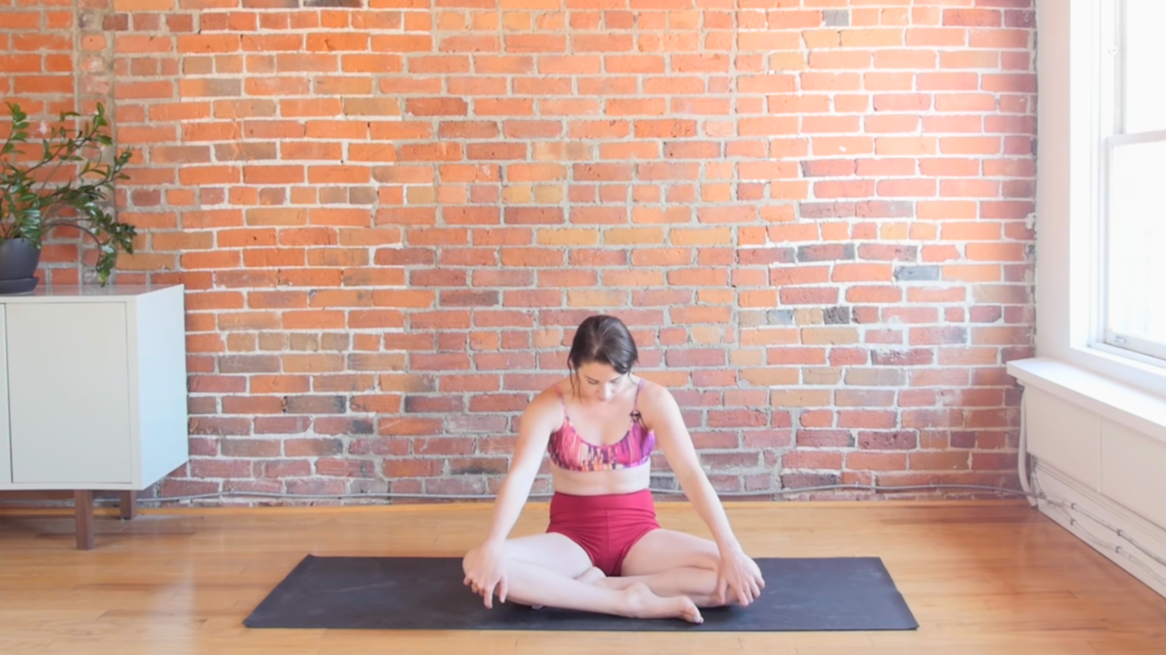 woman in seated cat pose on yoga mat in front of brick wall