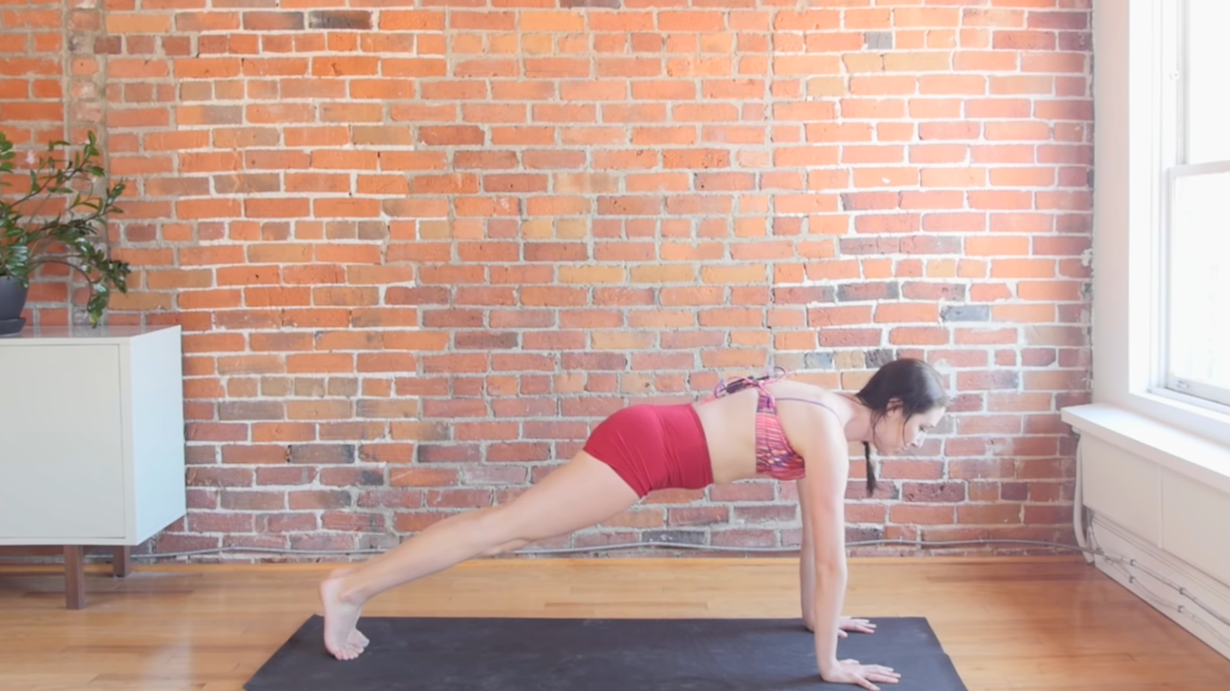 woman in plank pose over black yoga mat in front of brick wall