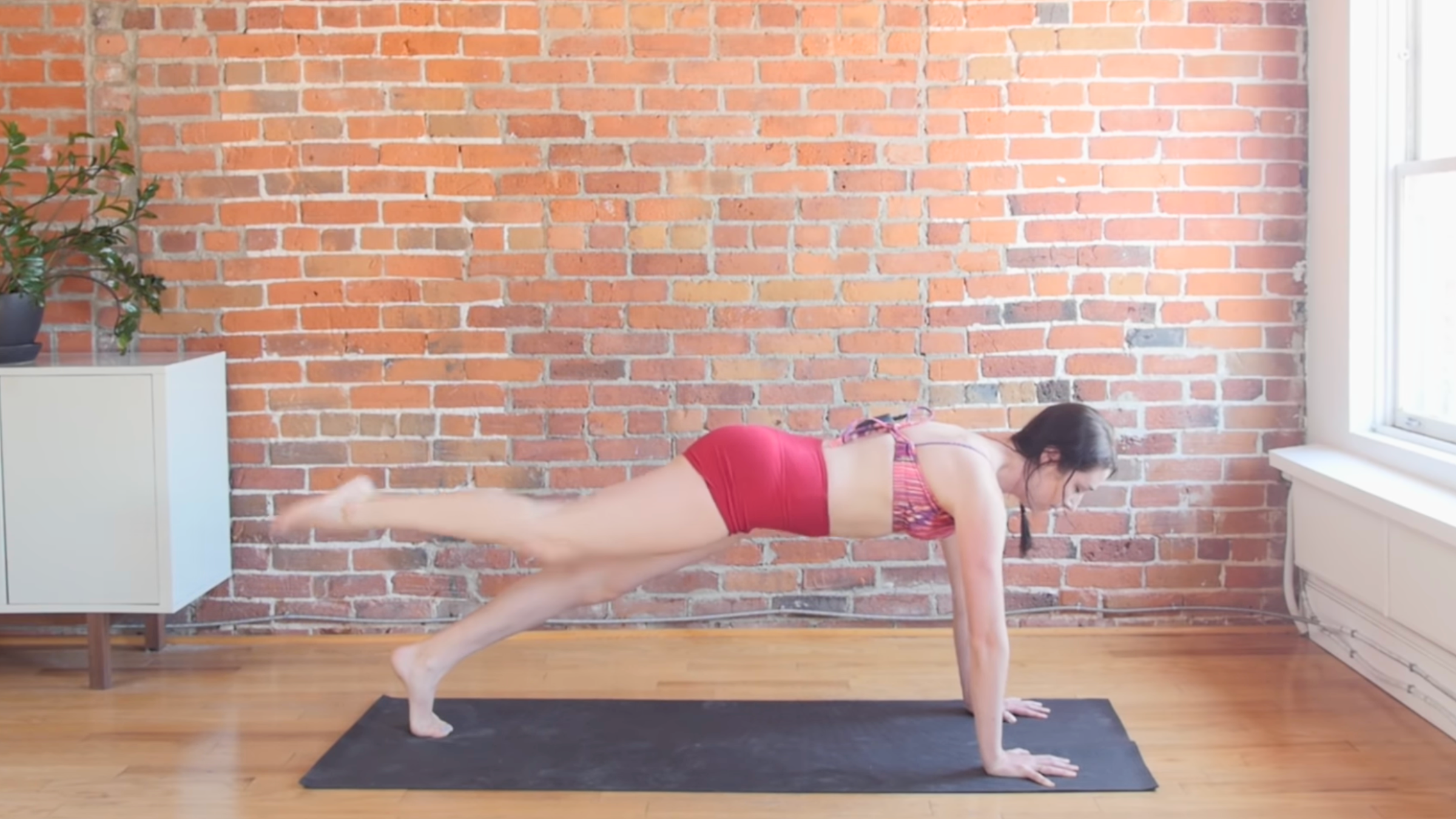 woman in plank pose with right leg lifted and parallel to the floor on black yoga mat in front of brick wall