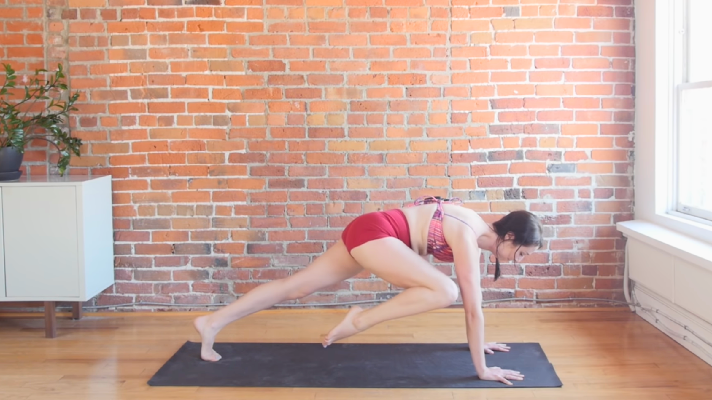 woman in straight arm plank pose with right knee touching right elbow on top of black yoga mat in front of brick wall
