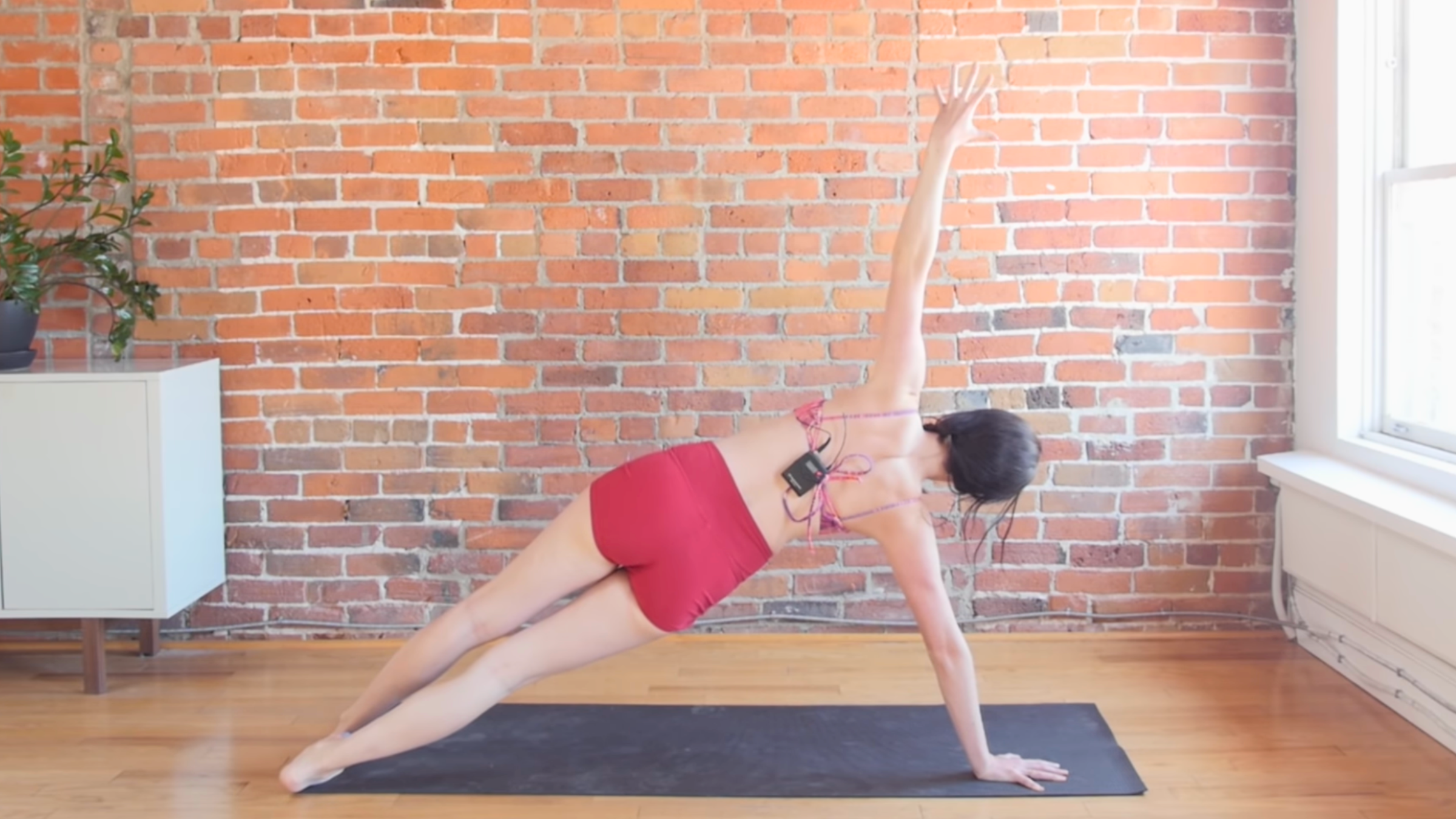 woman in side plank pose with back to camera on black yoga mat in front of brick wall