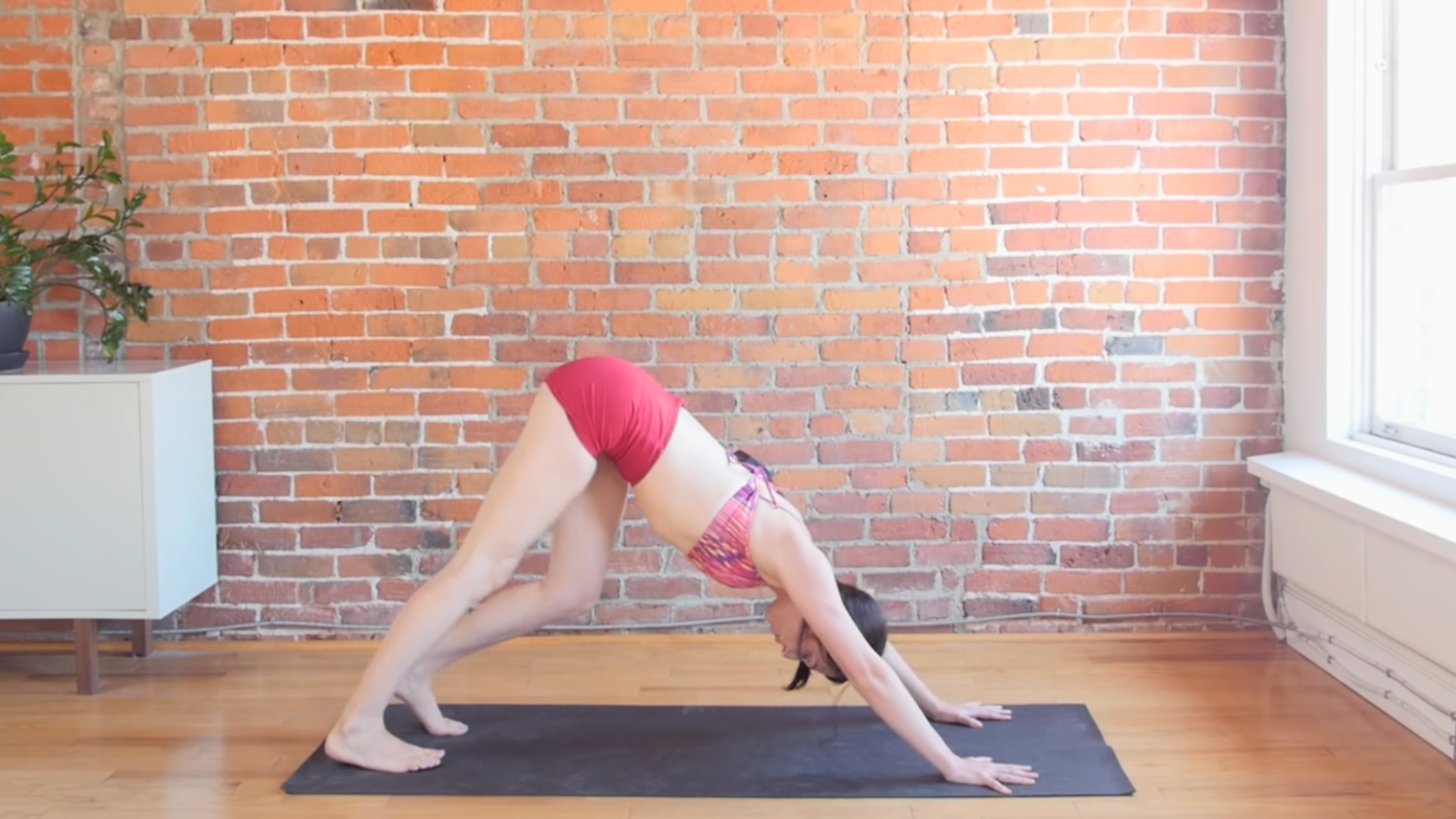 woman in downward dog pose while paddling her left foot on black yoga mat in front of brick wall