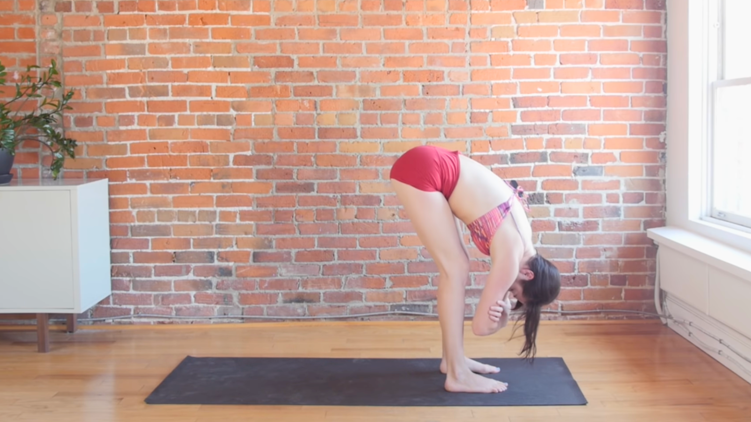 woman in standing forward bend pose with hands grabbing elbows on top of black yoga mat in front of brick wall