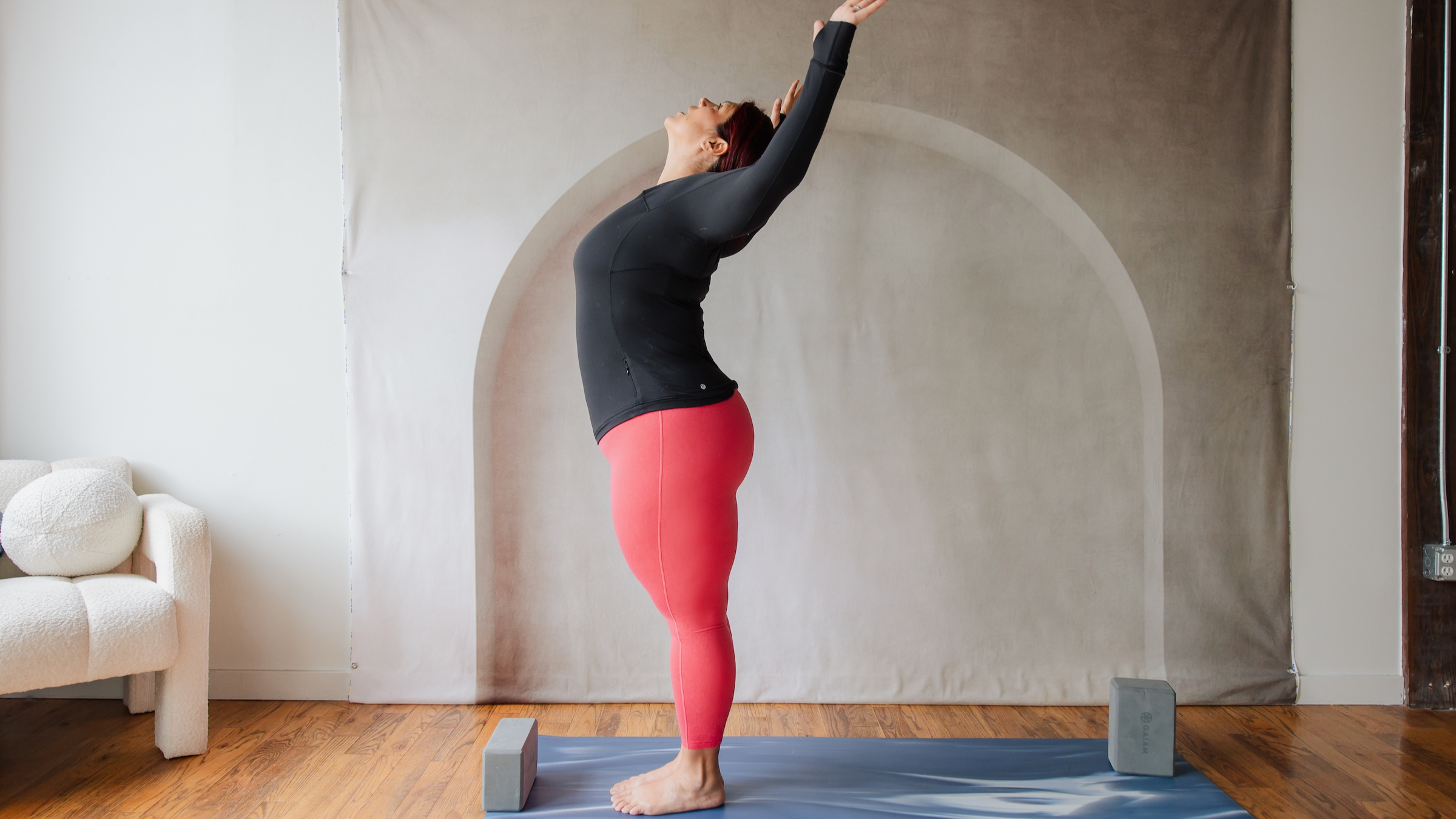 woman in black shirt and red yoga pants stands at the top of her yoga mat with her arms out in 90-degree angle cactus arms and face lifted toward the ceiling