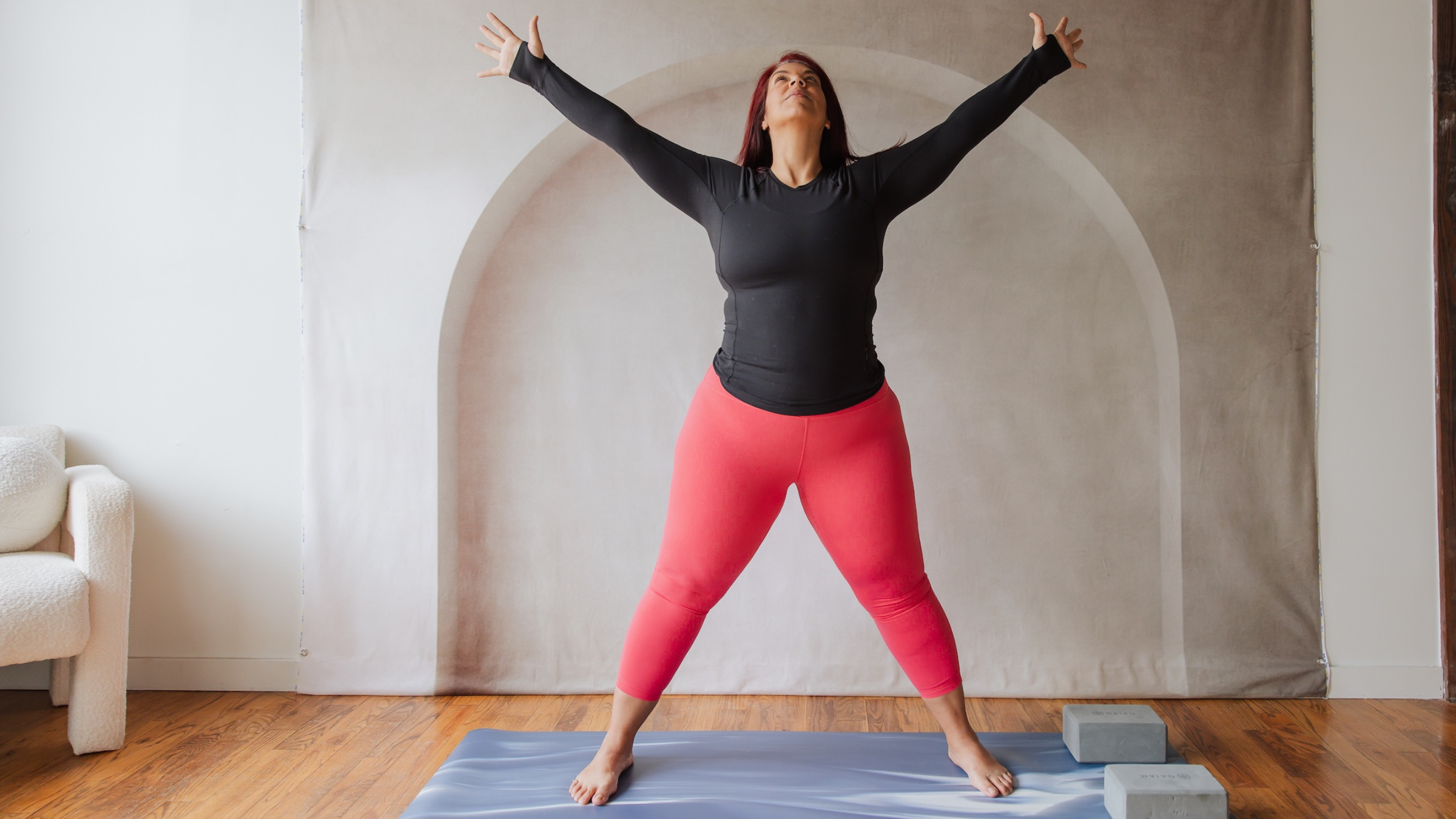 woman in black shirts and red yoga pants standing on yoga mat in star pose with feet out wide and arms extended
