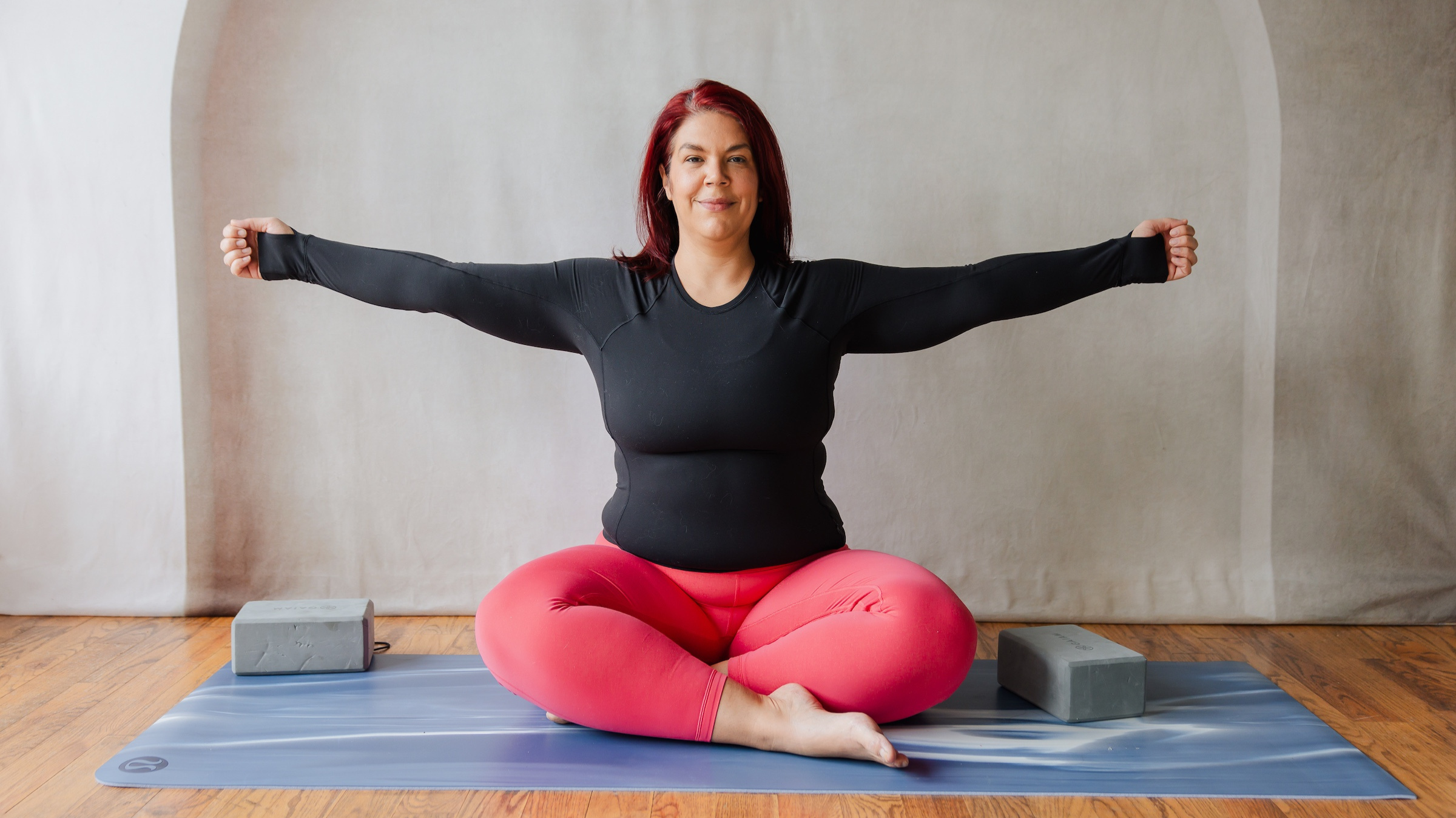 woman sits cross-legged on yoga mat on wooden floor with arms spread out to sides in black shirt and red yoga pants