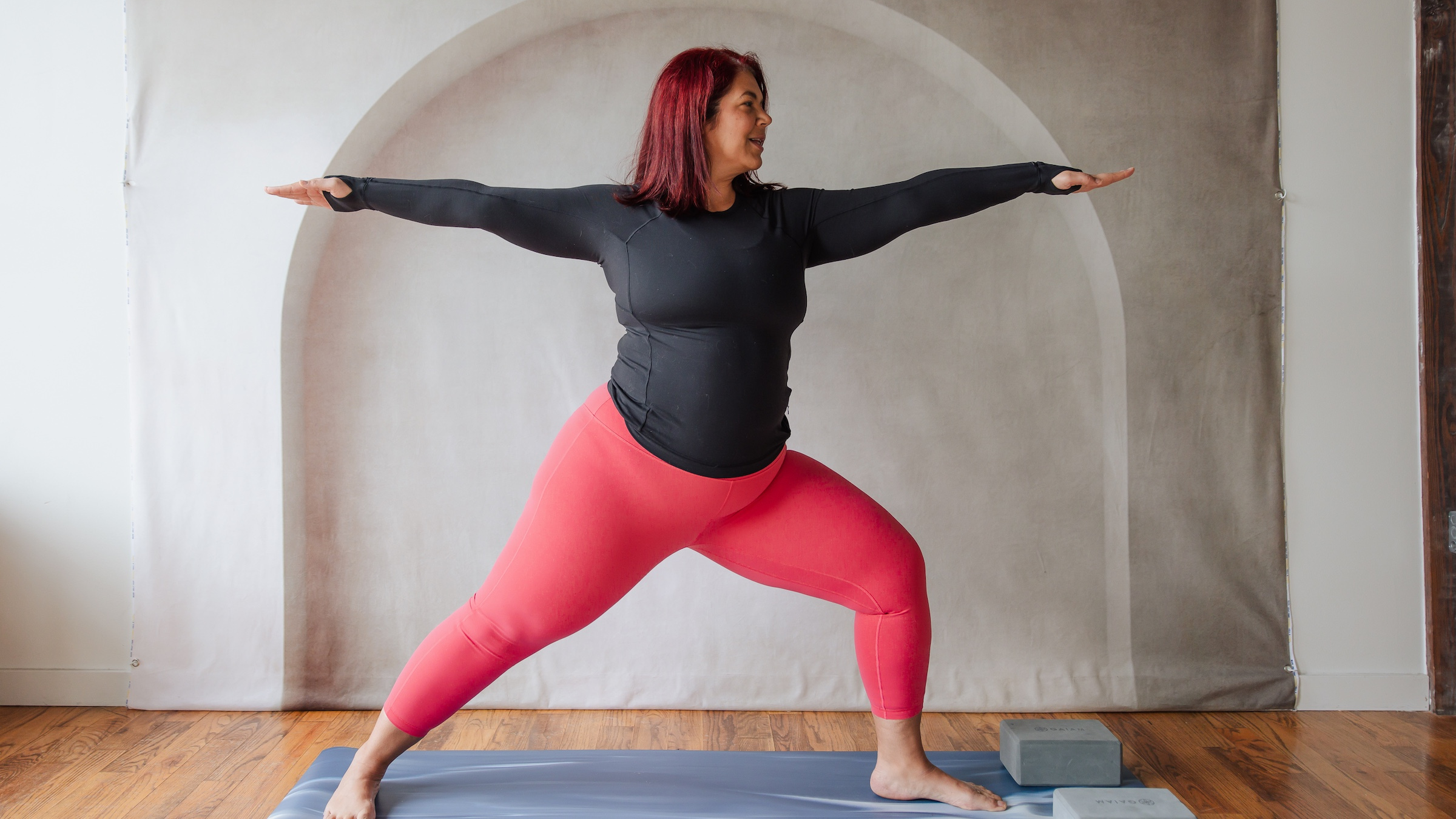 woman in black shirt and red yoga pants in warrior 2 pose with left foot bent at front of mat and arms extended parallel to the floor forward and backward