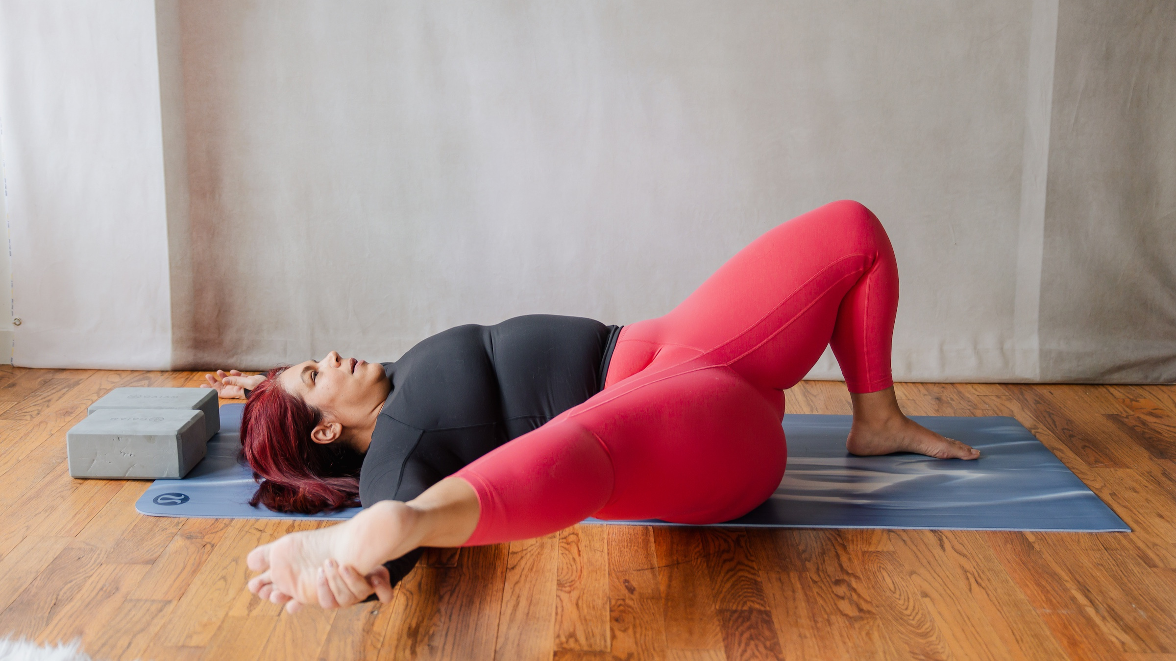 woman in black shirt and red yoga pants on back with left leg bent with foot on floor and right hand grabbing extended right leg