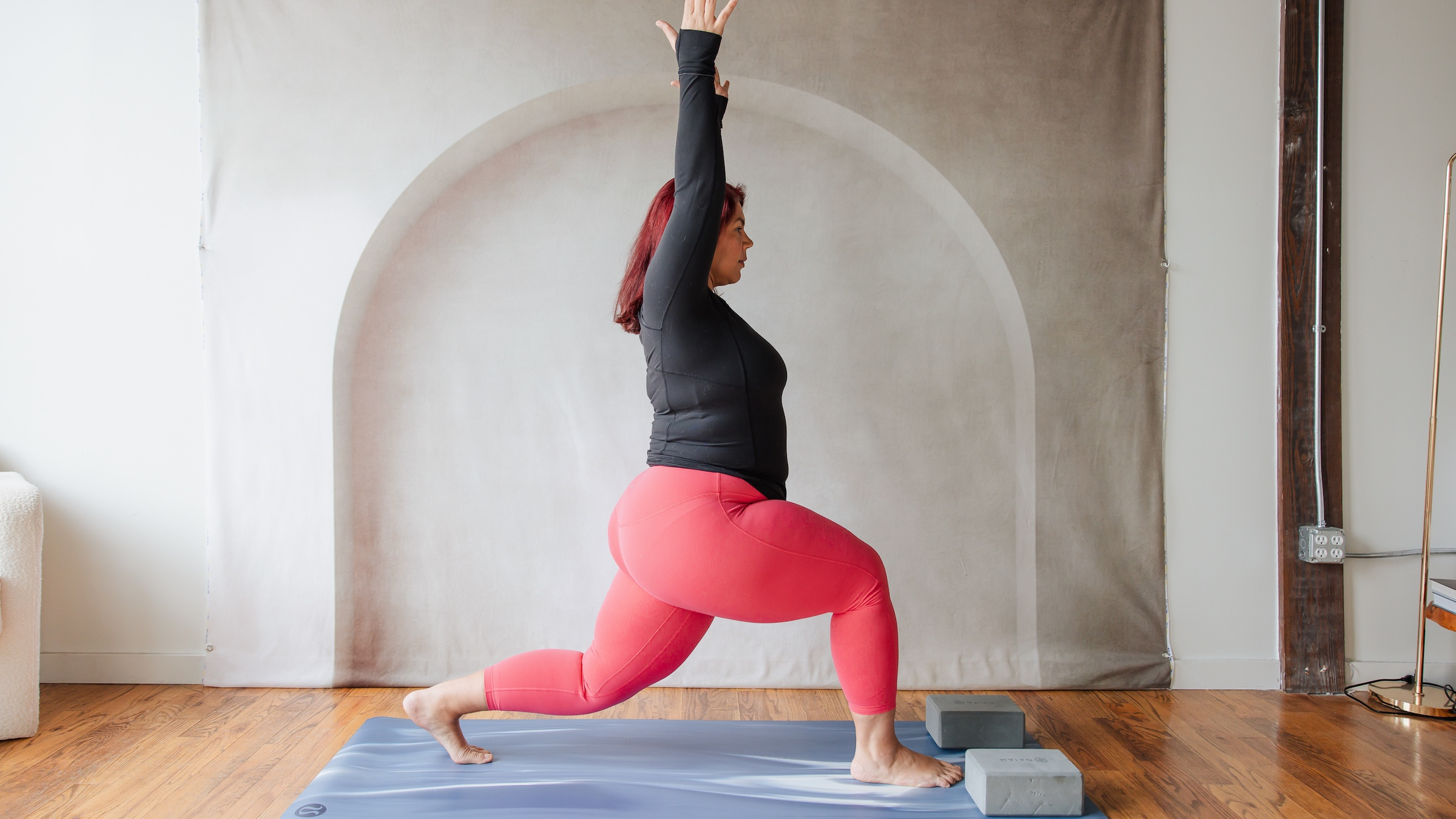 woman in black shirt and red yoga in standing lunge with arms above her head on yoga mat on top of wooden floor