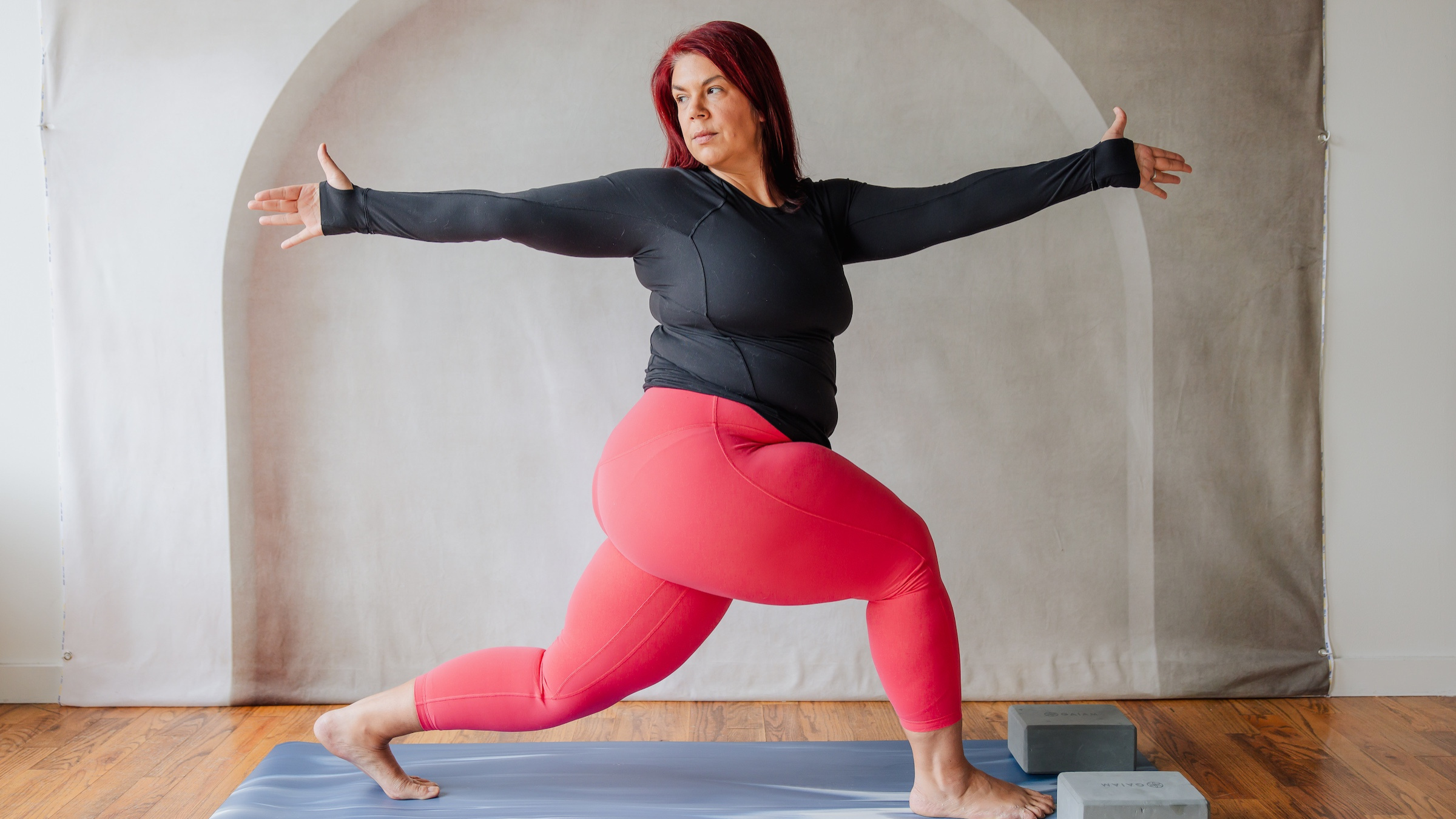 woman in black shirt and red yoga pants stands with legs in high lunge with upper body twisted the opposite way and arms extended forward and backward on yoga mat on wooden floor