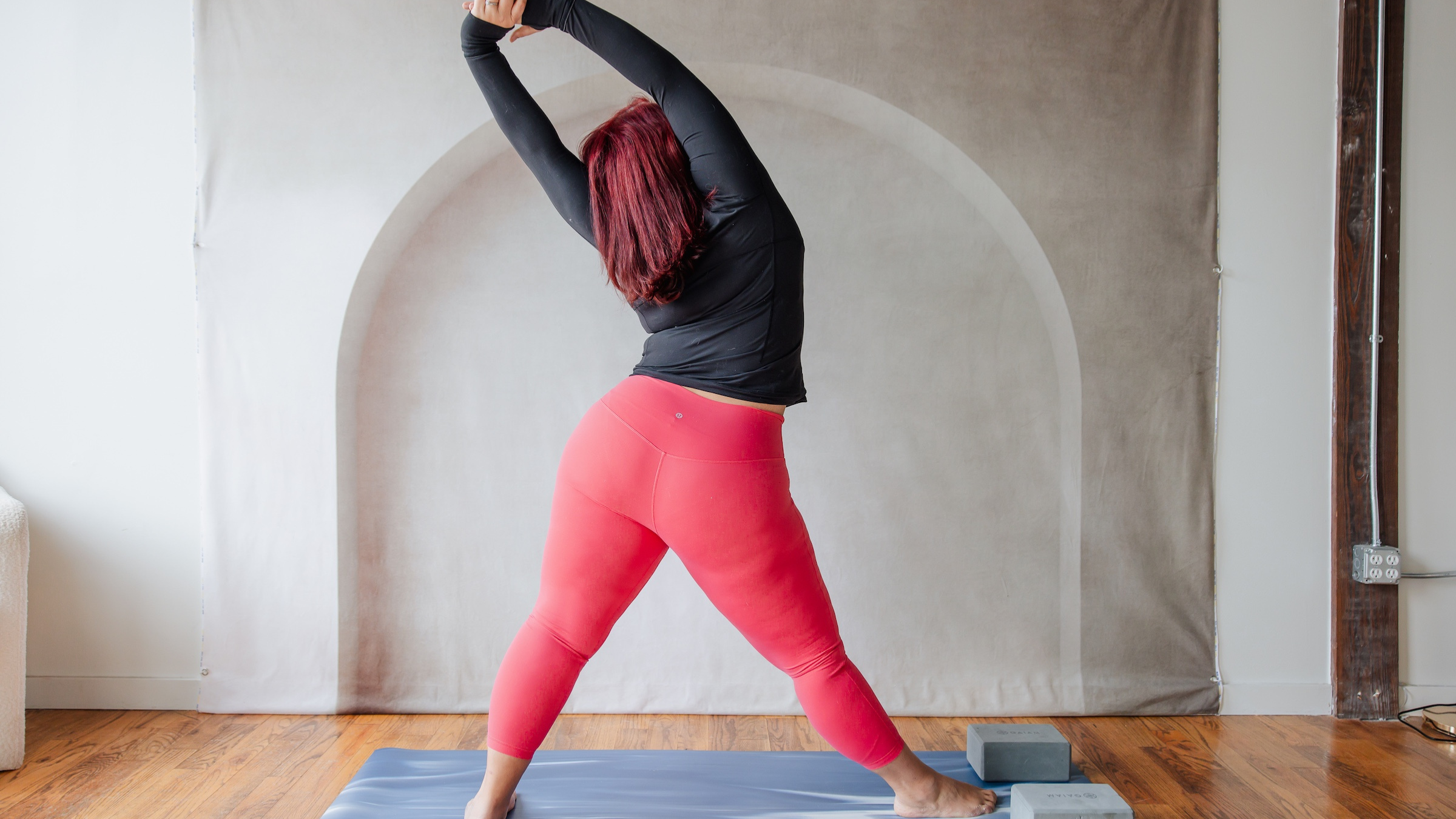 woman in black shirt and red yoga pants with legs in triangle pose with right foot forward while hands are clasped above head and are folded over to the left side of the body