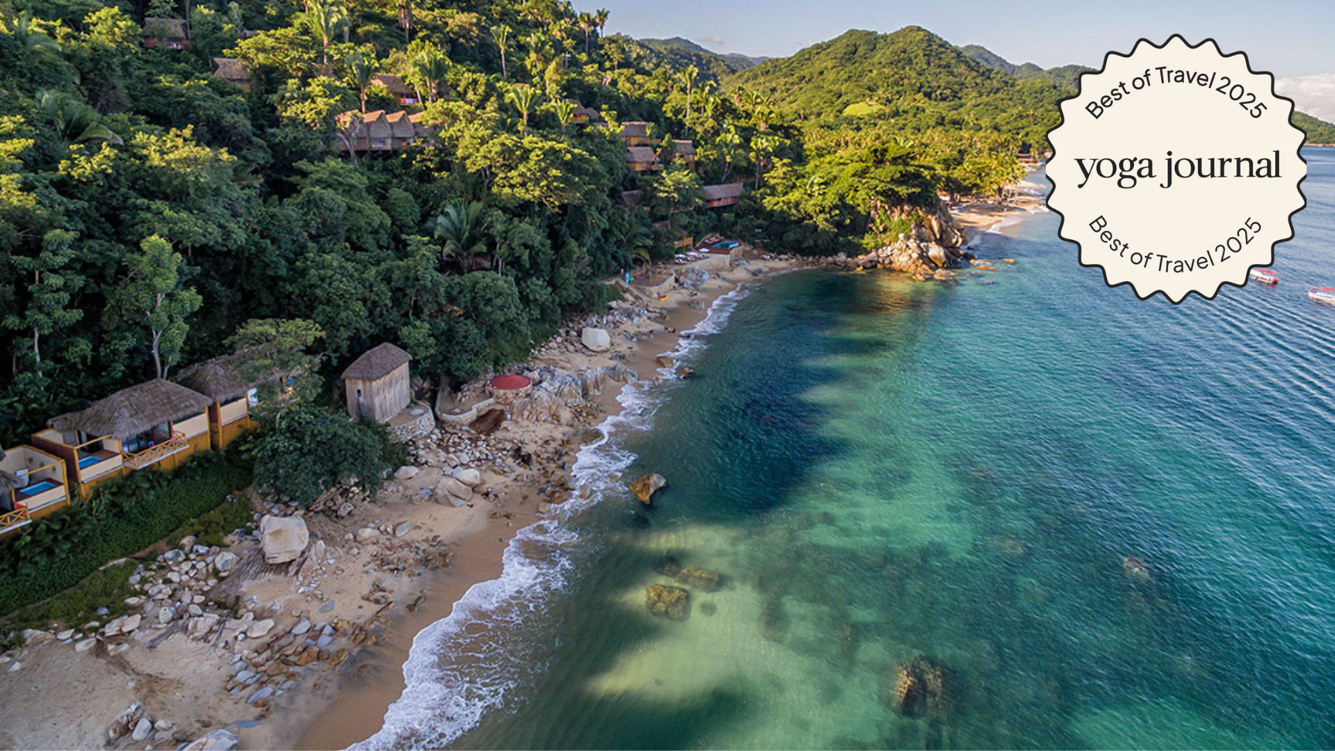 an aerial shot of a coastal yoga retreat center on the water with a forested hillside behind it