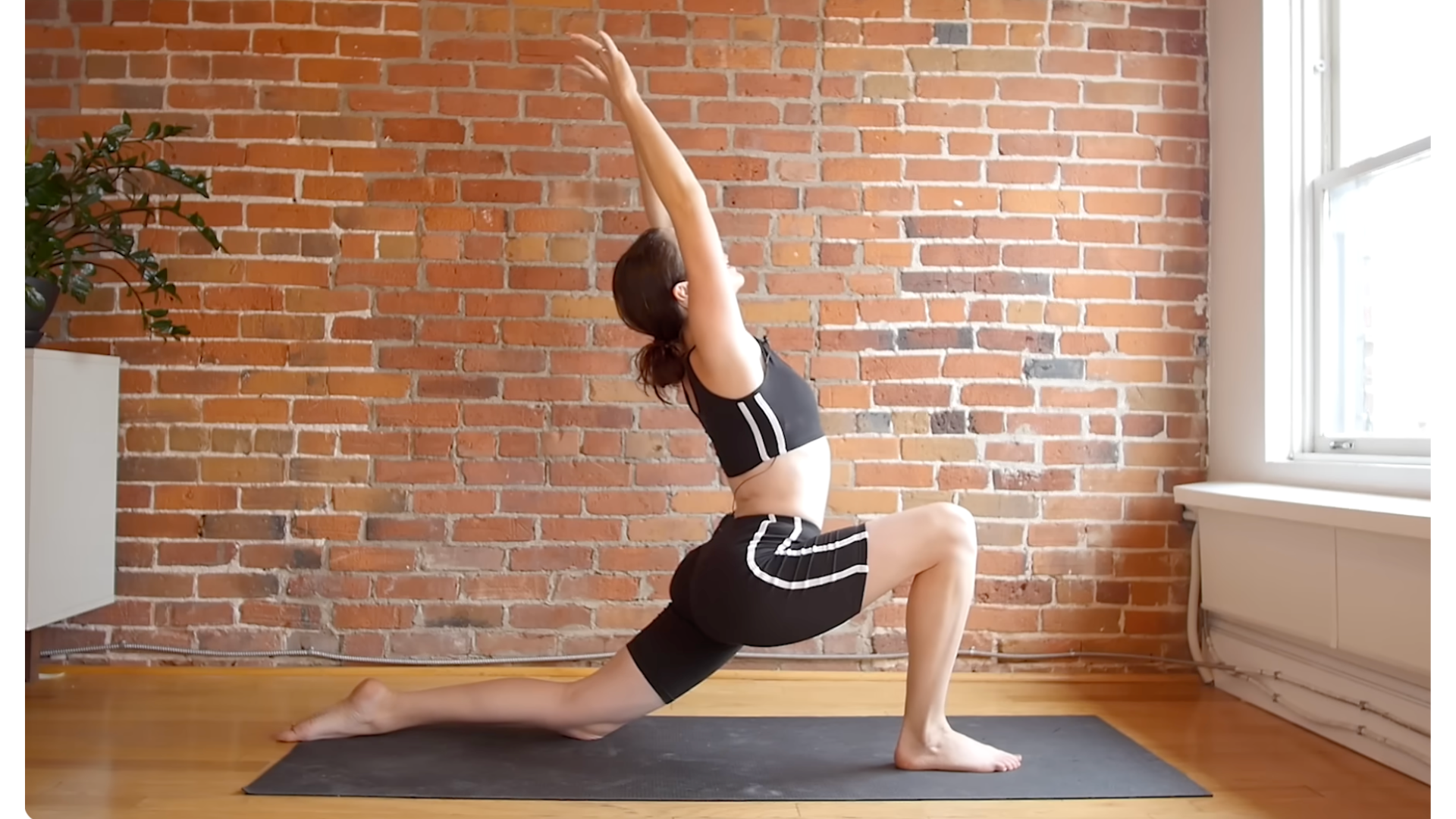 Yoga teacher on a yoga mat practicing a low lunge with her arms alongside her ears and her right foot forward for a stretch in her left hip flexors
