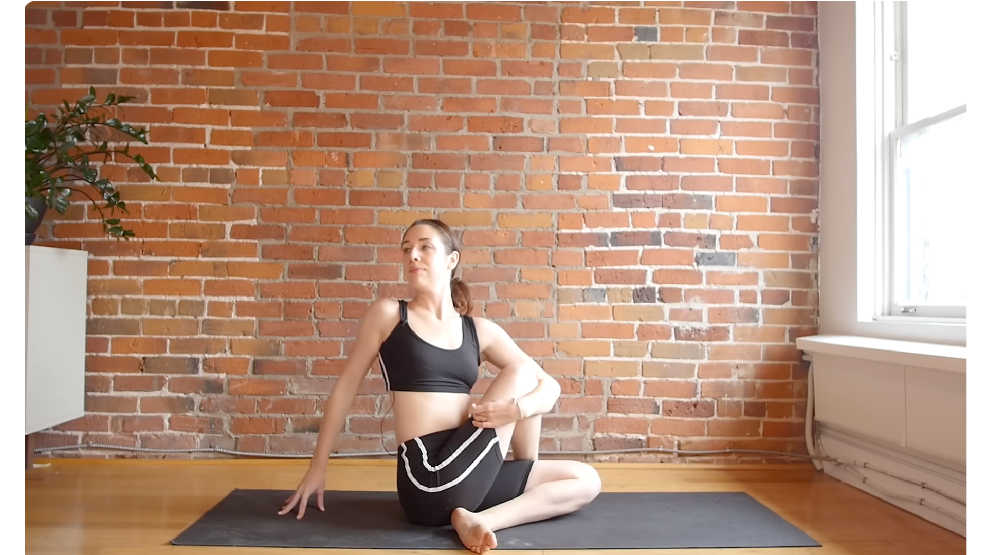 Yoga teacher sitting on a mat with both knees bent as she twists toward her right in a full-body stretch