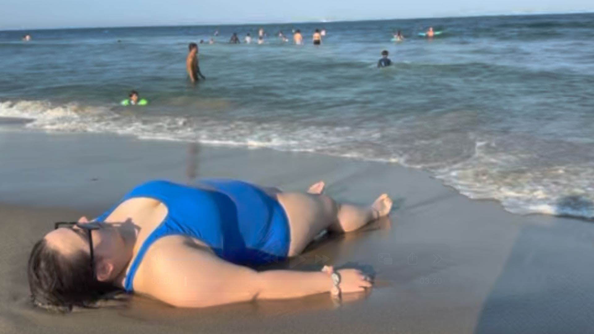 Image of woman in blue bathing suit lying down on wet sand near the ocean. People are standing in the water in the background.