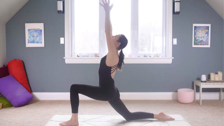 woman in black yoga clothes in Low Lunge Pose with arms extended straight overhead and face toward the ceiling on tan yoga mat on carpeted floor of home with window and blue wall behind her