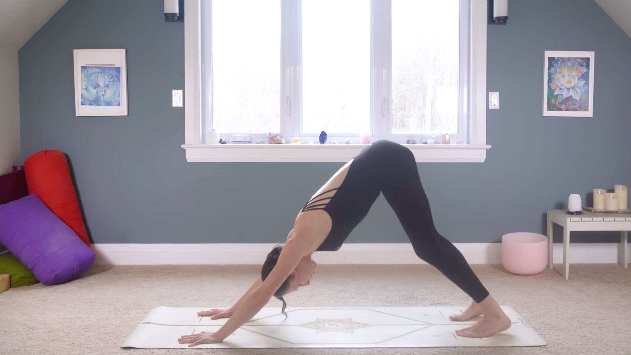 woman in black yoga clothes in Downward-Facing Dog Pose on tan yoga mat in home with carpeted floor, yoga items in background in front of windows and blue painted wall