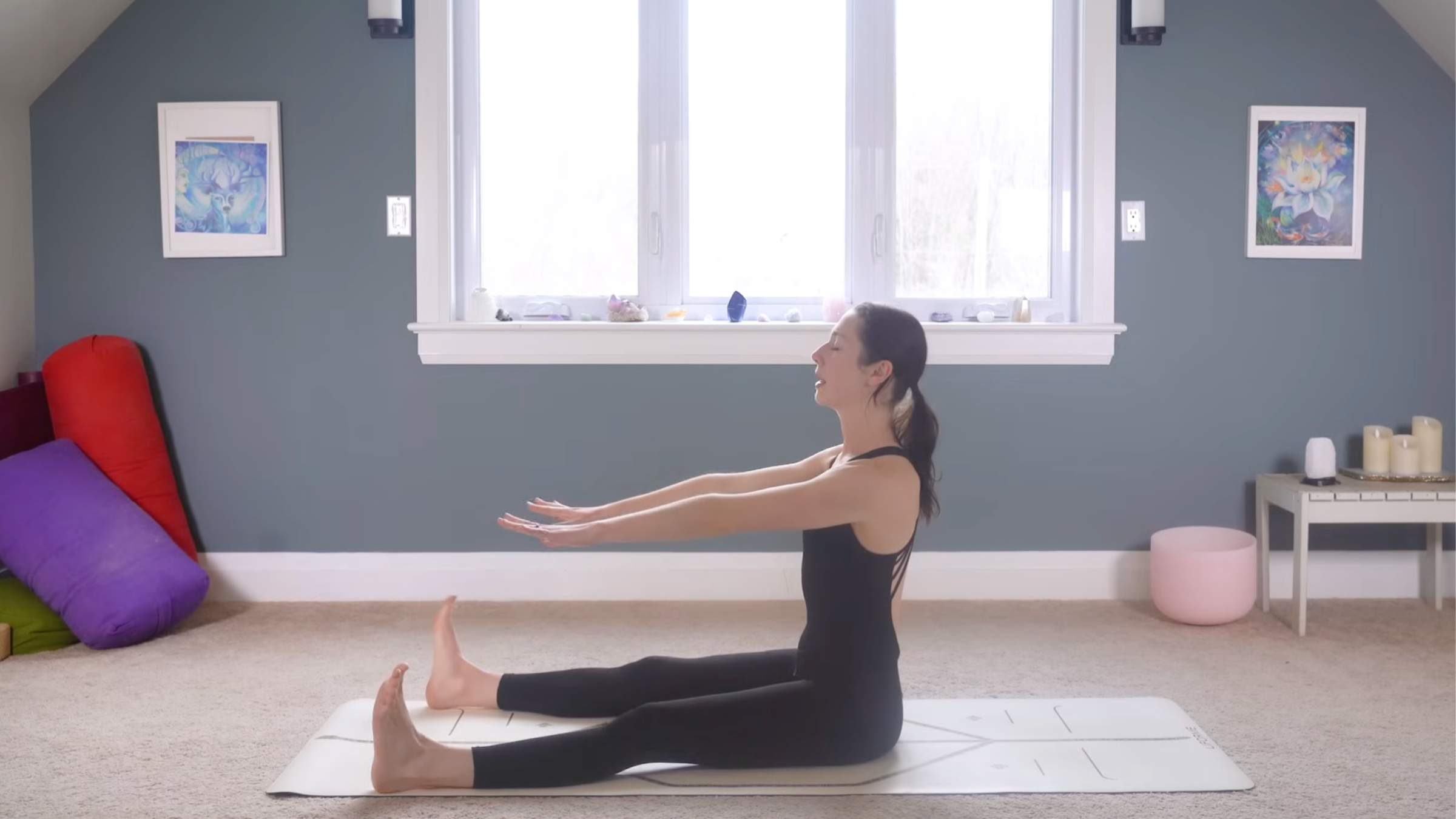 woman in black yoga clothes preparing for Spine Stretch Forward Pose by sitting with straight back, legs extended, feet flexed and arms extended toward feet on tan yoga mat in home with carpeted floor, yoga items in background in front of windows and blue painted wall