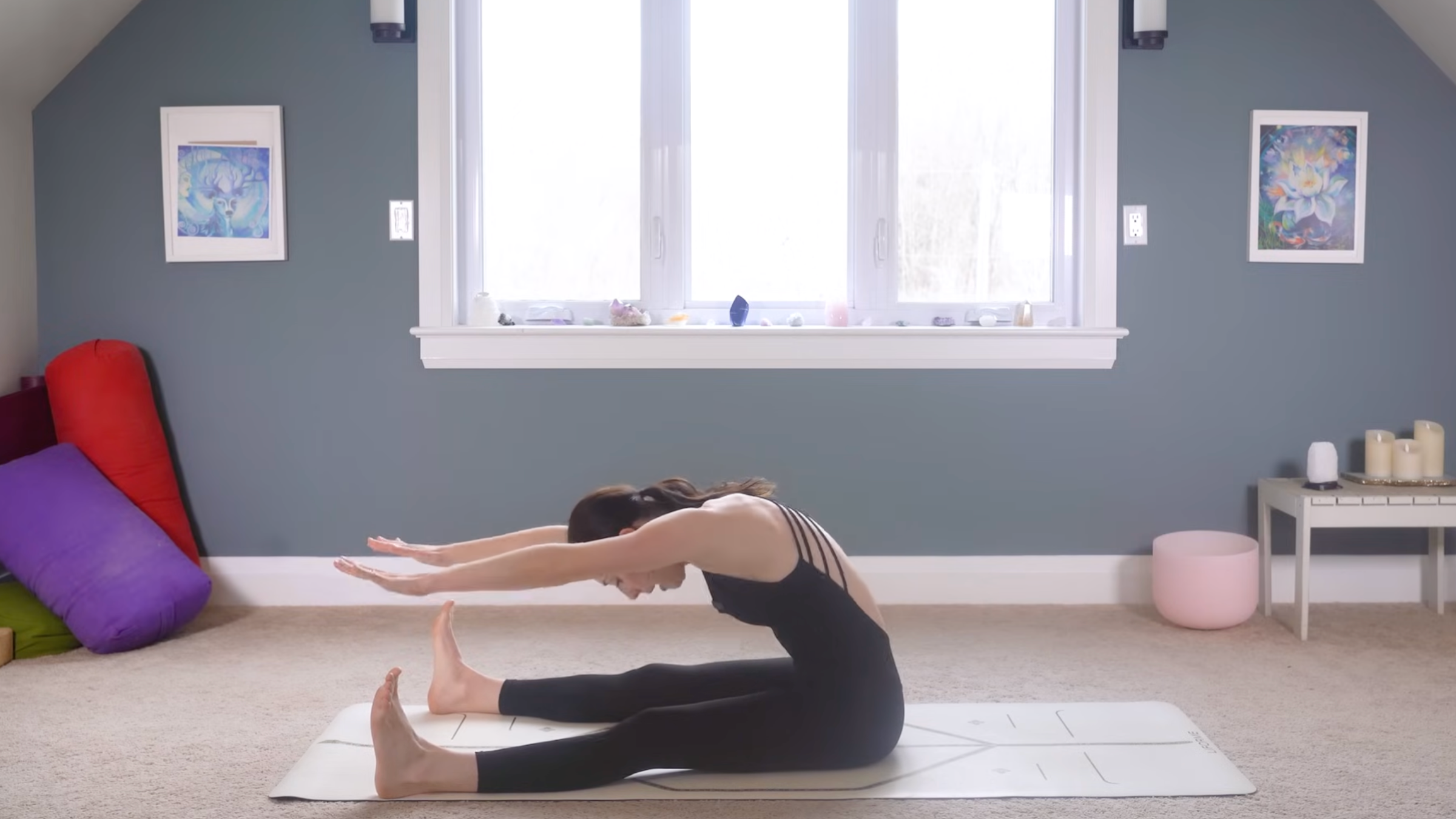 woman in black yoga clothes stretching spine with legs extended on ground, feet flexed, and spine curved with arms reaching forward on tan yoga mat in home with carpeted floor, yoga items in background in front of windows and blue painted wall