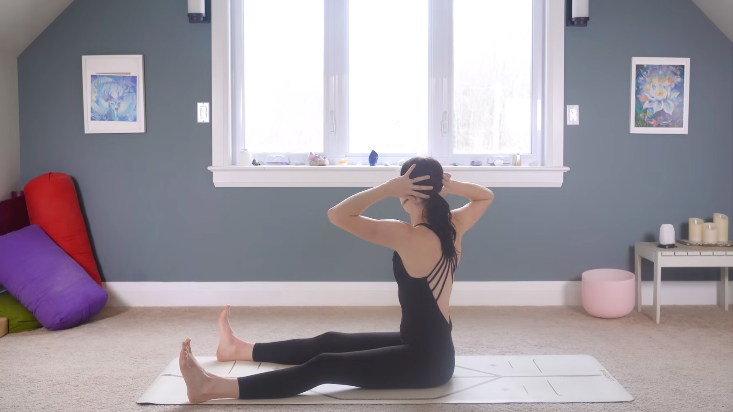 woman in black yoga clothes in seat spine twist with legs extended on ground, feet flexed, arms on sides of head and torso twisted toward right side on tan yoga mat in home with carpeted floor, yoga items in background in front of windows and blue painted wall