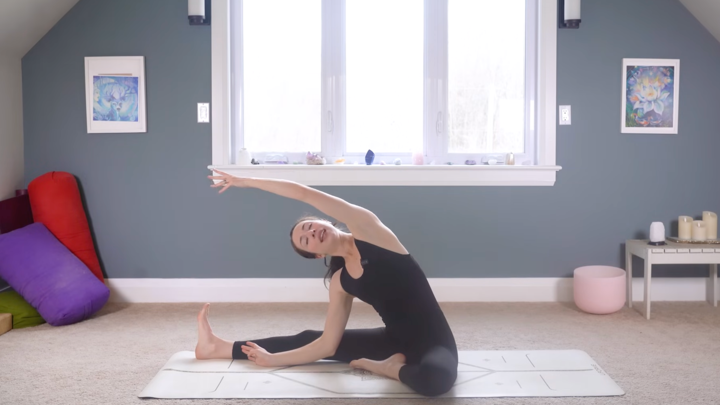woman in black yoga clothes stretching side body with right leg extended on ground, left leg bent, and left arm reaching up and overhead toward left foot on tan yoga mat in home with carpeted floor, yoga items in background in front of windows and blue painted wall