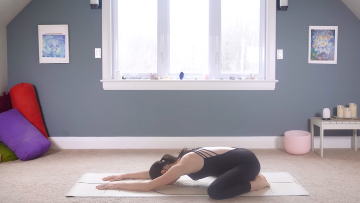 woman in black yoga clothes in Child's Pose on tan yoga mat on carpeted floor of home with window and blue wall behind her