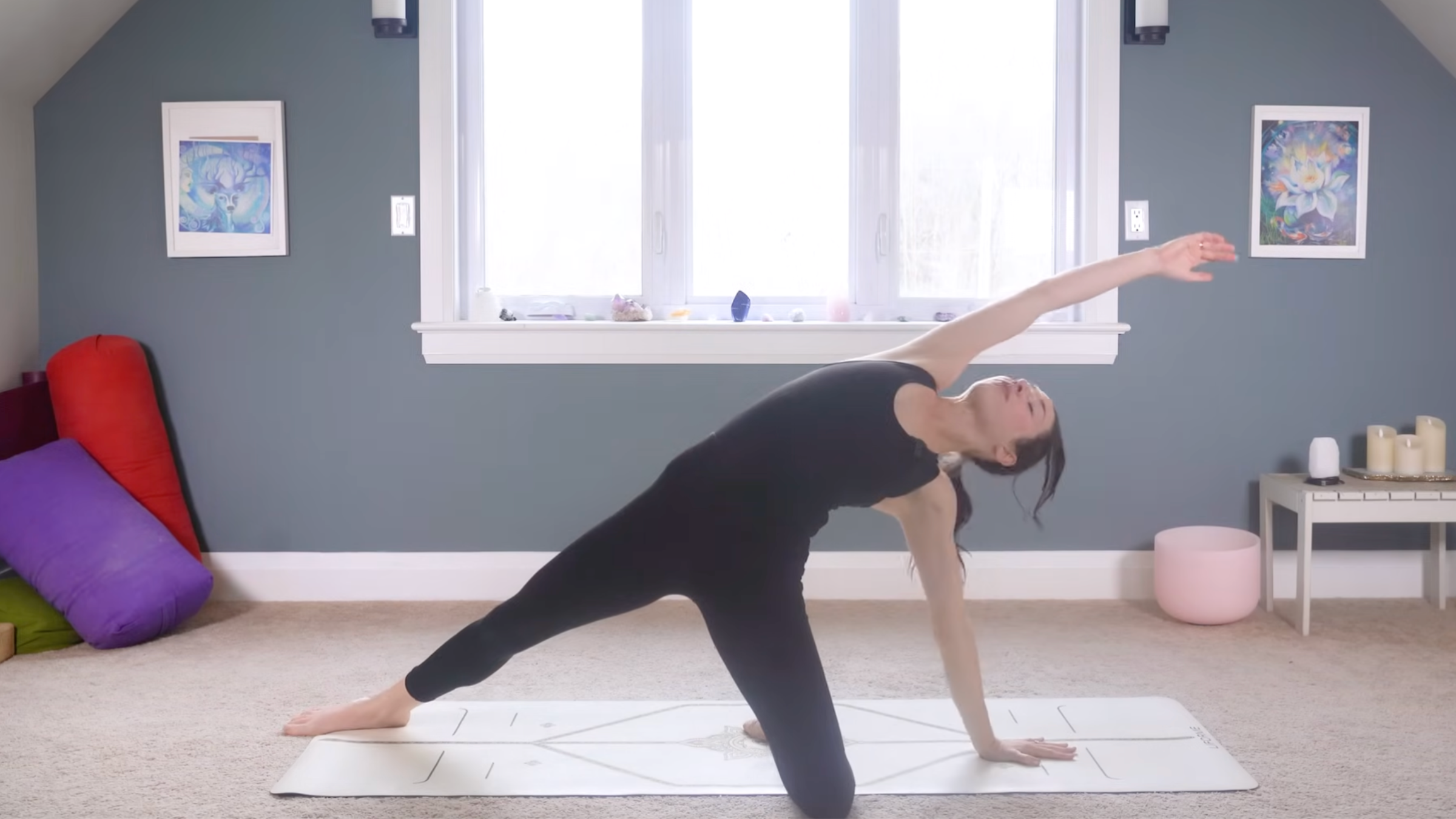 woman in black yoga clothes in Baby Wild Thing Pose with left knee, shin, and hand on the ground, right leg extended, and right arm extended overhead on tan yoga mat in home with carpeted floor, yoga items in background in front of windows and blue painted wall
