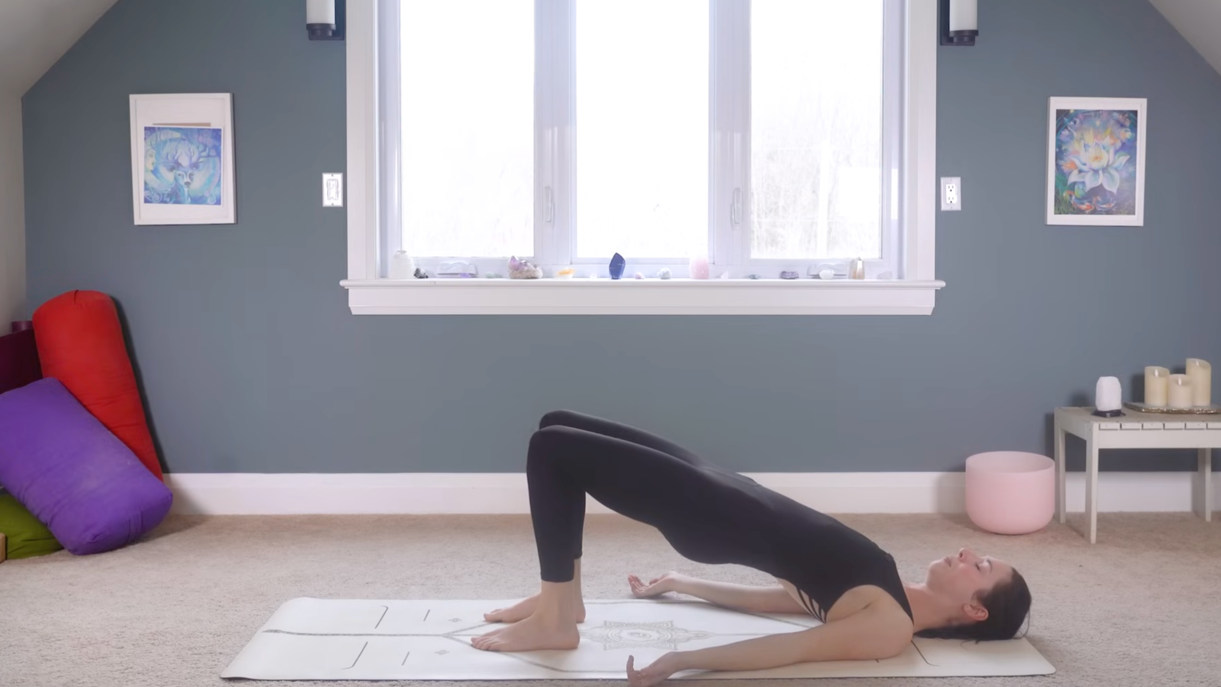 woman in black yoga clothes in Bridge Pose with arms, feet, shoulders and head on the ground, and hips elevated for a straight spine on tan yoga mat in home with carpeted floor, yoga items in background in front of windows and blue painted wall