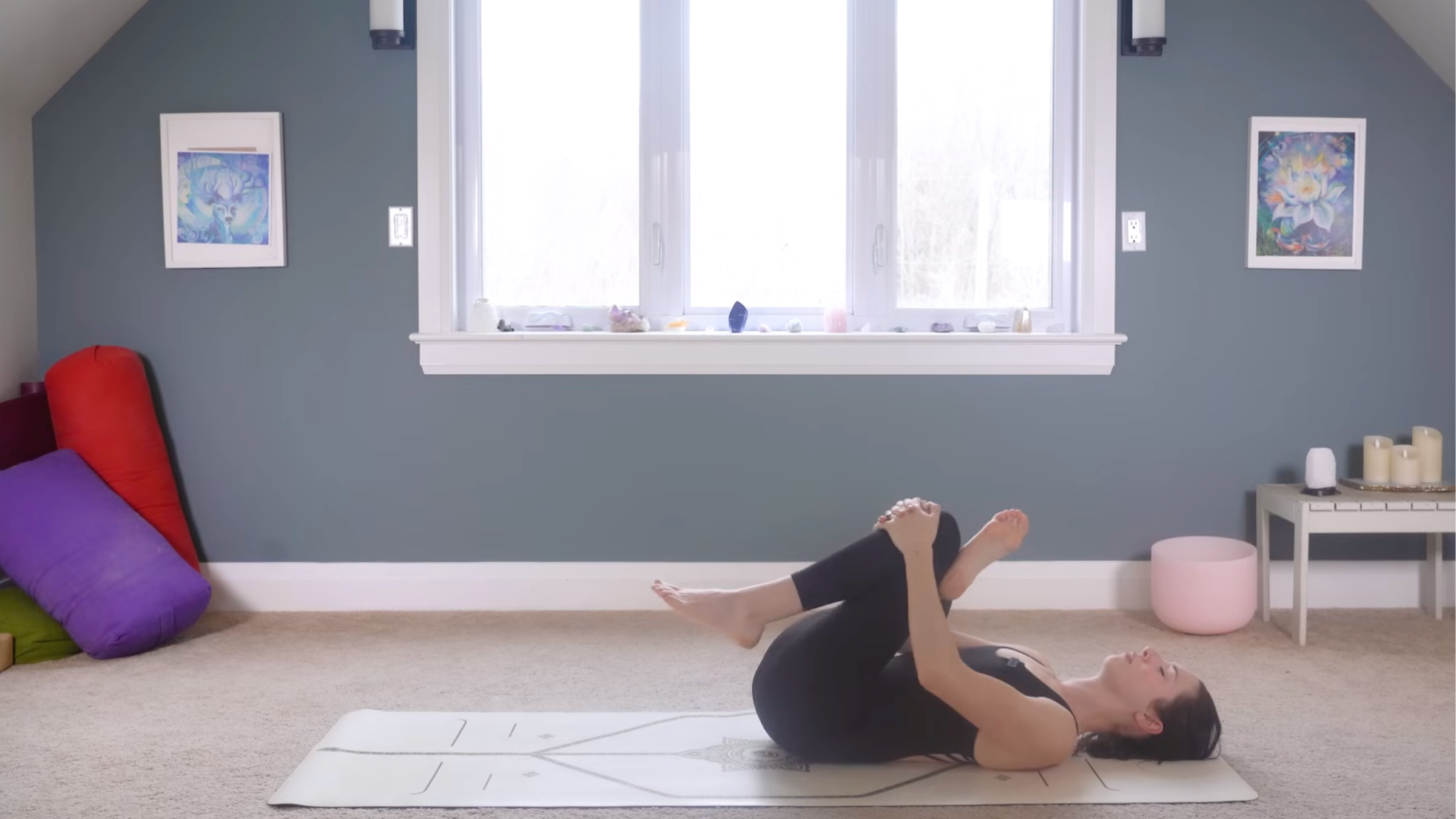 woman in black yoga clothes in reclined pigeon pose lying down right right ankle crossed over right thigh, both knees bent, and legs grabbing left knee, on tan yoga mat in home with carpeted floor, yoga items in background in front of windows and blue painted wall