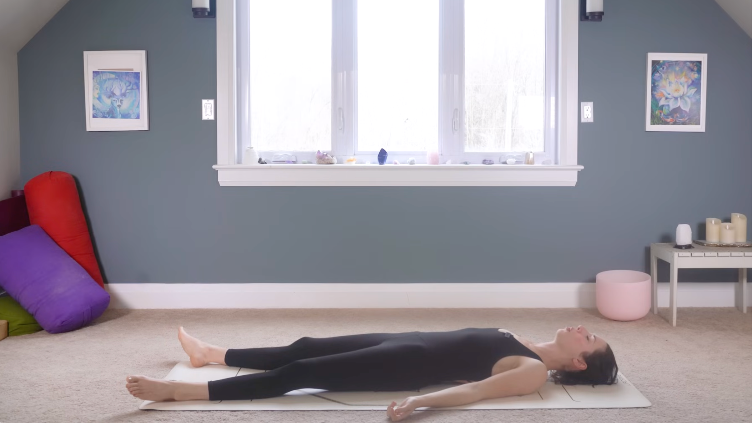 woman in black yoga clothes in Savasana pose lying relaxed on tan yoga mat with legs and arms extended in home with carpeted floor, yoga items in background in front of windows and blue painted wall