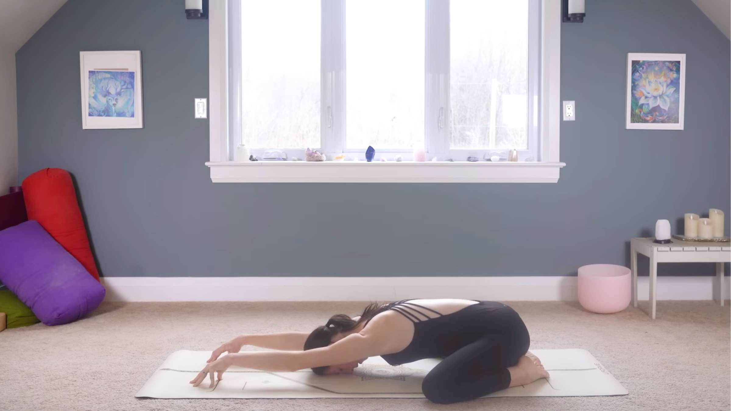 woman in black yoga clothes in Child's Pose but with wrists and elbows elevated and only fingertips touching the mat, on tan yoga mat on carpeted floor of home with window and blue wall behind her