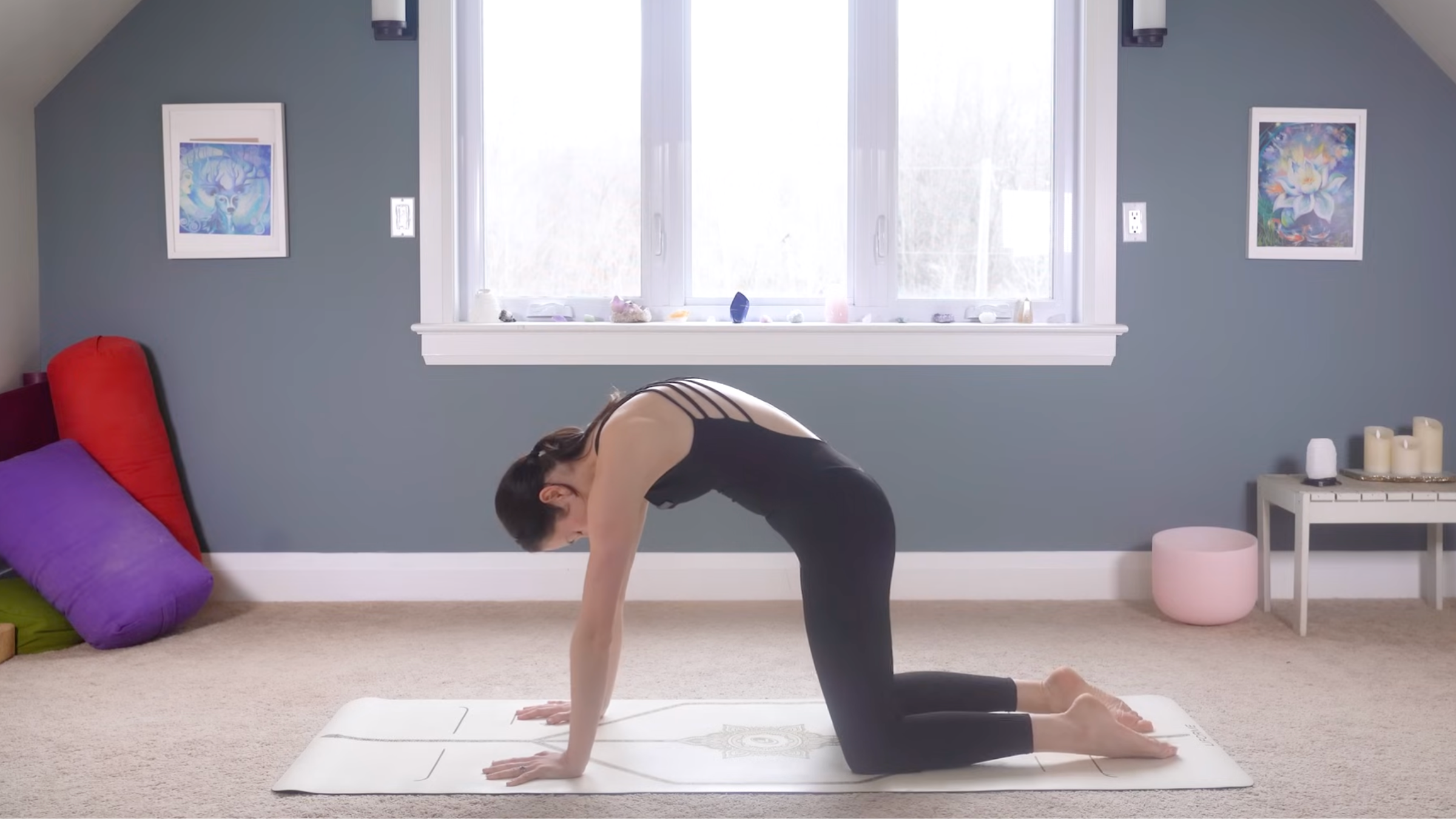 woman in black yoga clothes in Cat Pose on tan yoga mat on carpeted floor of home with window and blue wall behind her